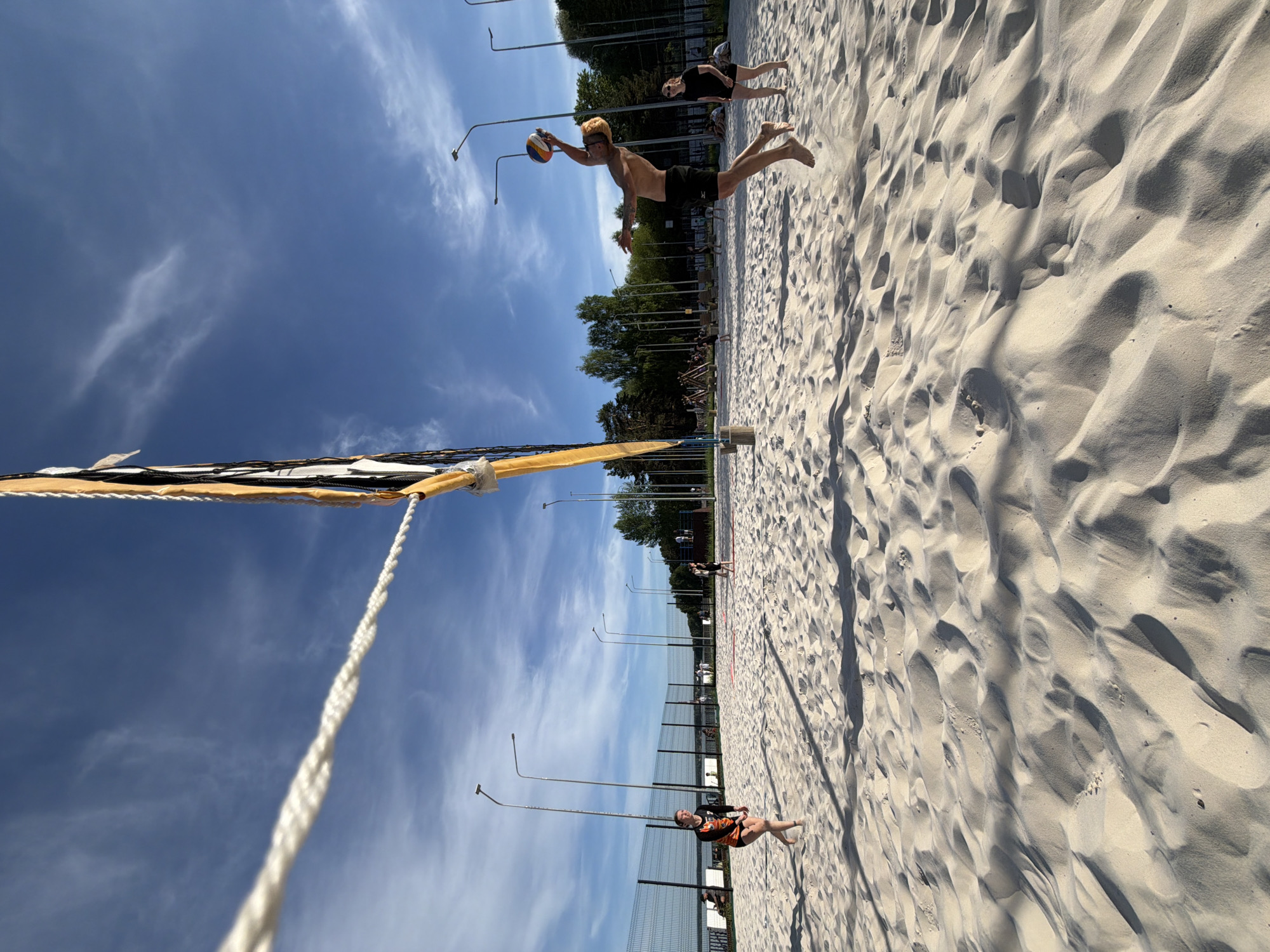 The image shows a beach volleyball game in progress on a sunny day. A man is diving to hit the ball near the net, while another person is lying on the sand in the background. In the foreground, a person is lying on the ground closer to the net, also appearing to be playing volleyball. The beach is sandy, and there are trees and lampposts in the background. The location is Chaikovsky, Russia.