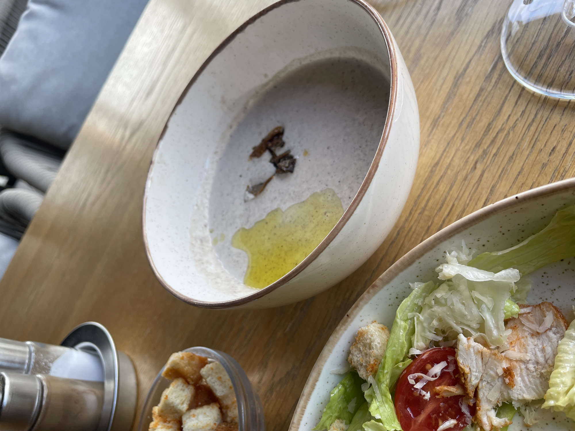 The image shows a close-up of a table setting at a restaurant, likely in Sochi, Russia, given the context of the provided information. On a wooden table, there is a bowl of mushroom soup with a swirl of olive oil and some pieces of mushroom garnish. To the left of the soup bowl is a partially visible serving of chicken salad, featuring lettuce, grilled chicken breast, tomato, and croutons. A small glass bowl filled with croutons is also present. In the background, a salt and pepper shaker set sits on a metallic base. The environment appears to be an indoor dining area with soft lighting, possibly with other diners or furniture visible out of focus in the background.