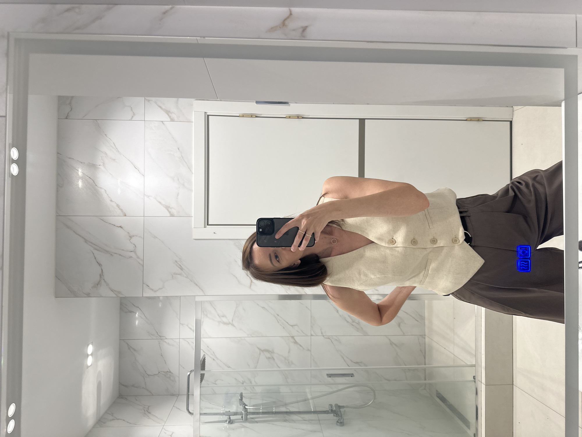 A woman is standing in front of a mirror in a modern bathroom, taking a selfie with her phone. She is wearing a light beige vest over a light-colored necklace and dark brown wide-leg pants. The bathroom features marble-patterned tiles on the walls, a glass-enclosed shower, and a white door. The mirror has a built-in light strip at the top and displays a small blue icon of two squares with wavy lines inside them, possibly indicating a digital control or display. The location is not specified as Прикамский, Russia; this information is not present in the image.