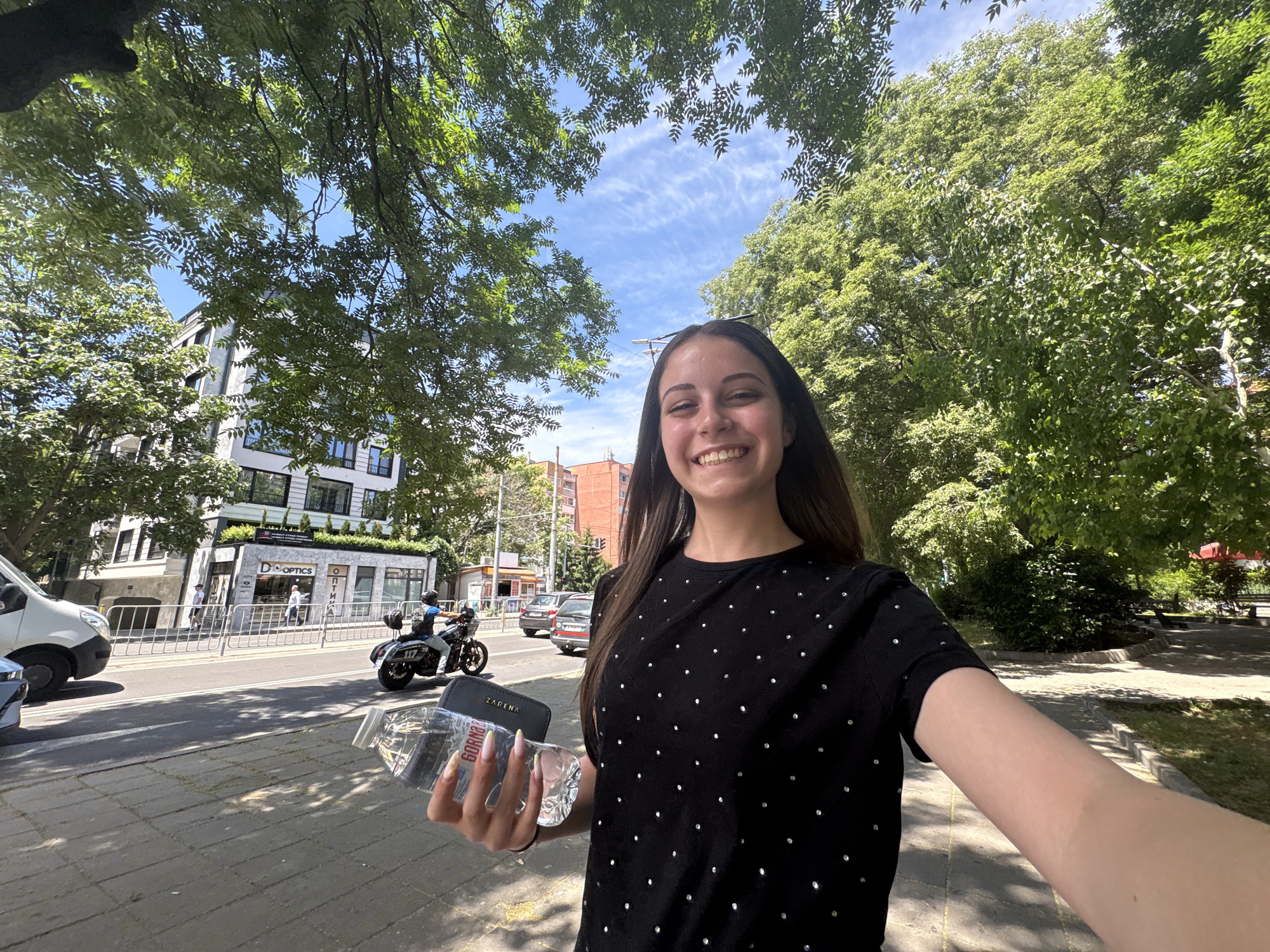 A young woman with long, dark hair and a bright smile takes a selfie outdoors on a sunny day. She is wearing a black t-shirt adorned with scattered white dots. In her right hand, she holds a clear plastic bottle of water and a black wallet with "ZARENA" embossed on it.
The background shows a sidewalk next to a street with some traffic. A white van is parked on the left, and a police motorcycle with the number "117" on it is visible further down the street, with a police officer on board. In the distance, there's a building with shops, one labeled "D'OPTICS". Trees with lush green foliage frame the top of the image, suggesting a warm, pleasant weather. The sky is blue with scattered white clouds. The overall scene depicts a casual outdoor moment, possibly a walk in a city or town. The time of day appears to be late morning or early afternoon due to the bright sunlight and distinct shadows.