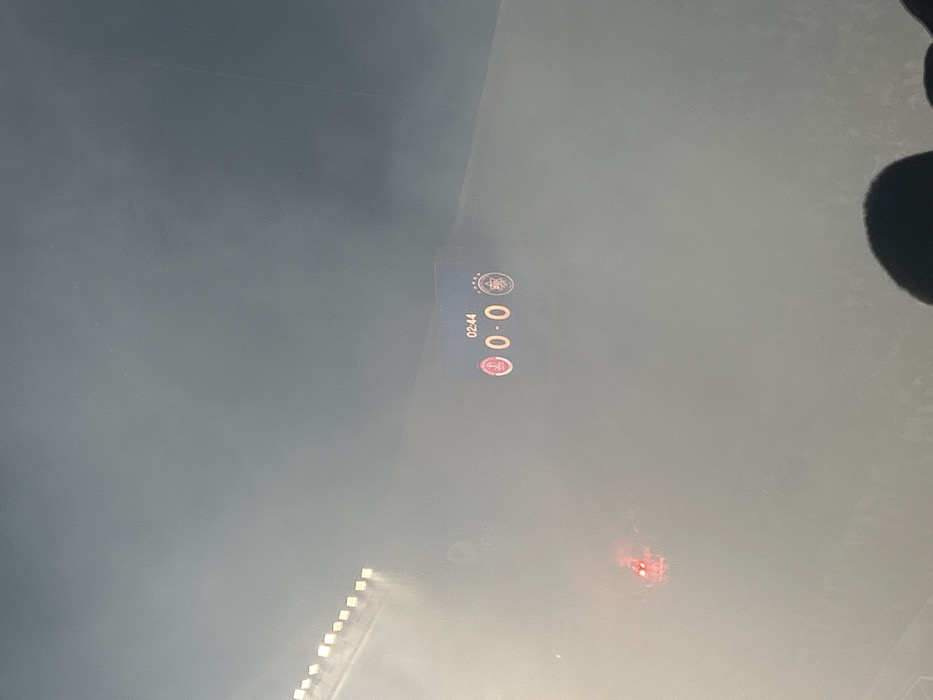 A blurry image shows a soccer scoreboard displaying the score 0-0 at the 02:44 mark. The scoreboard features team logos, one potentially red. A line of stadium lights is visible in the lower left, and a red light, likely a flare, is burning to the right. A dark blurry shape is visible in the top right corner of the image. The entire image is hazy, suggesting smoke or fog.