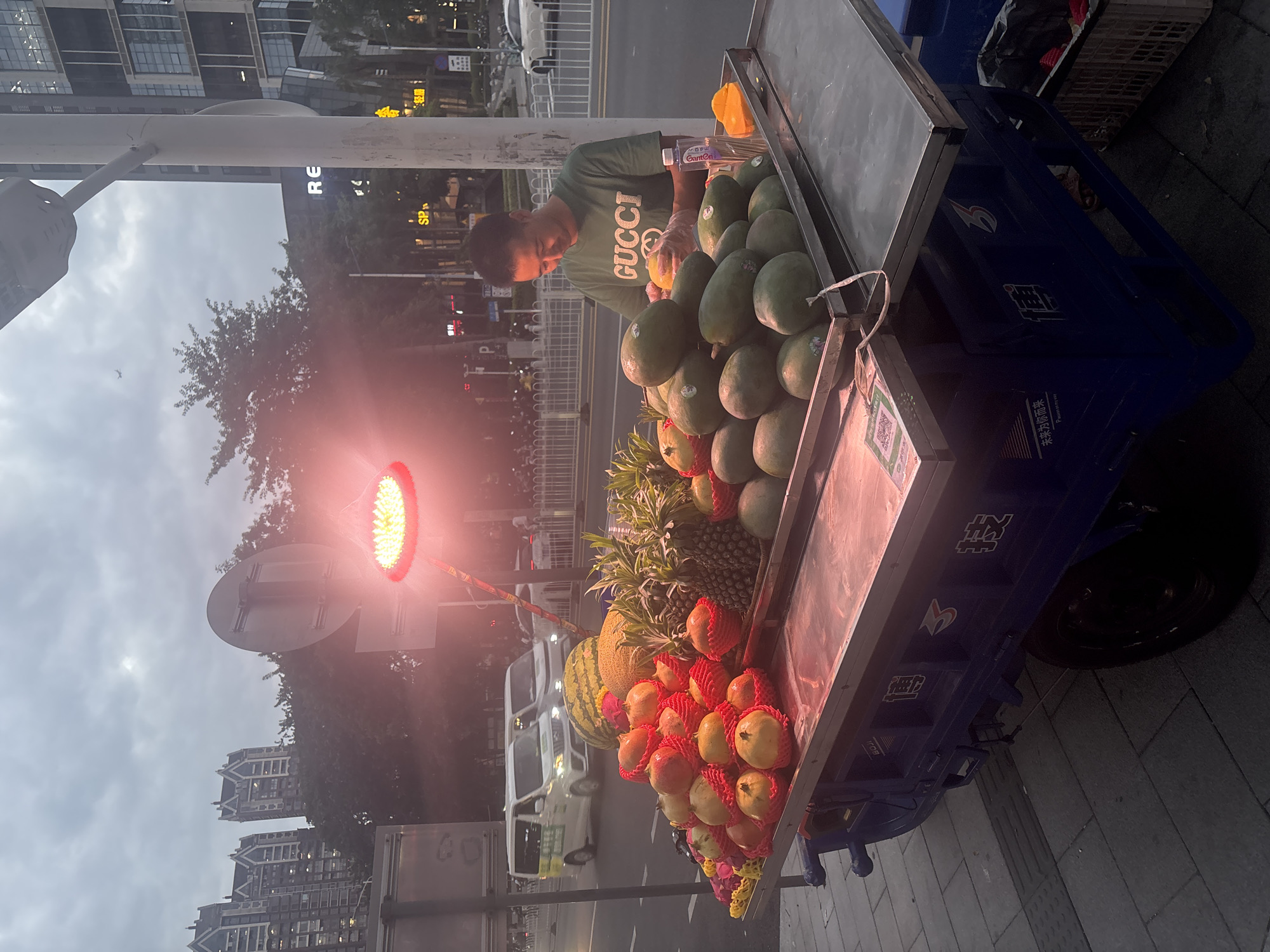 A man wearing a green t-shirt with "GUCCI" printed on it is standing behind a blue fruit cart. He is wearing transparent gloves and appears to be peeling a piece of yellow fruit. The cart is laden with various fruits, including pomegranates, pineapples, melons, and what appear to be papayas. A bright red light is illuminated above the cart, casting a reddish glow. In the background, there are tall buildings, trees, and vehicles on a street. The sky is overcast. The overall scene suggests a street-side fruit vendor in an urban environment, possibly in Liwan District, China, given the style of the buildings and the script on the cart. The time of day appears to be dusk or early evening.