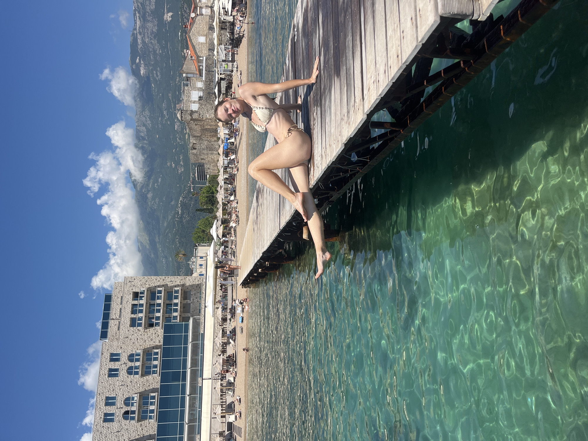 A woman in a light-colored bikini sits on a wooden pier extending over clear, turquoise water. She is leaning back on her right hand, with her left arm bent and her left leg extended. The pier is made of light-colored wooden planks. Beneath the pier, dark metal supports are visible. The water is calm with visible ripples and light reflections.
In the background, a sandy beach is lined with lounge chairs and umbrellas. Beyond the beach, there is a modern building with large windows and stone cladding, followed by older stone buildings with terracotta roofs. Mountains rise in the distance, partly obscured by clouds. The sky is bright blue with scattered white clouds. The scene appears to be set during a sunny day, likely in the late morning or early afternoon, given the strong sunlight and clear sky. The woman's pose suggests relaxation and enjoyment of the beach or vacation setting. The location is identified as Budva, Montenegro. No visible text is present in the image.