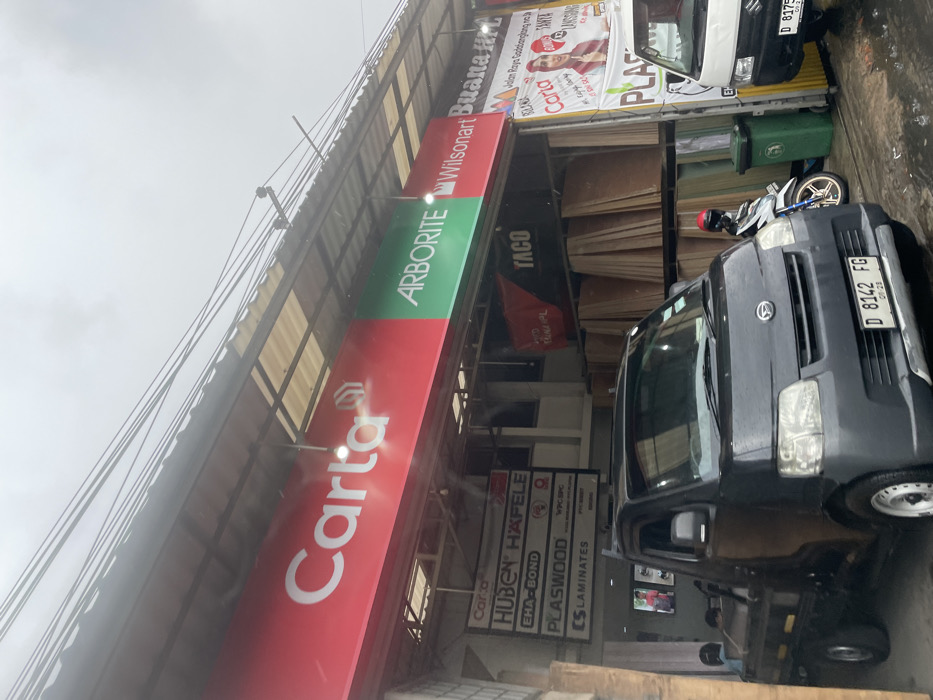 The image shows a storefront in Ngamprah, Indonesia, located at coordinates -6.868, 107.521. The storefront has a large red sign with the word "Carta" in white letters. To the right of the sign is a green sign with the words "Arborite," "Wilsanart," and "Buana" in white letters. The storefront appears to be a building materials store. There are various stacks of plywood behind the signs. A black pickup truck is parked in front of the storefront. The pickup truck has a license plate that reads "D 8142 FG." To the right of the pickup truck, there is a motorcycle and a green trash can. Another white pickup truck can be seen in the right-hand corner of the image. The truck has a partially obscured license plate that reads "D 8179." The storefront is located on a street with a grey asphalt surface. There are a few wires overhead. The weather is overcast, and the street is wet. A person wearing a red helmet can be seen in the reflection of the pickup truck's window. In the background, a person can be seen walking towards the storefront.