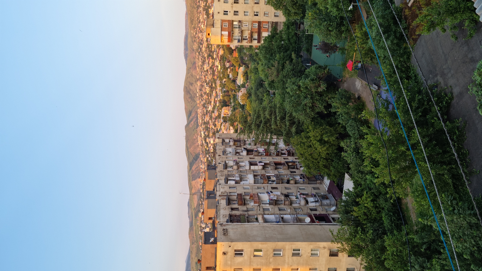 The image presents an aerial view of a residential area in Tbilisi, Georgia, likely during the golden hour. In the foreground, a weathered, multi-story apartment building dominates the left side, its balconies adorned with laundry and satellite dishes. Adjacent to it, another apartment building stands on the right, with similar architectural features. A dense canopy of green trees separates the buildings from a small recreational area below, which includes a playground with a red and yellow structure, a basketball court, and some scattered benches. Beyond the immediate residential structures and greenery, the city of Tbilisi unfolds in the background, a sprawling landscape of smaller buildings and houses stretching towards rolling, tree-covered hills that define the city's edge. The sky is a clear, pale blue, transitioning to a warmer hue near the horizon, suggesting either sunrise or sunset. The overall impression is a glimpse into the urban fabric of Tbilisi, characterized by its Soviet-era apartment blocks, abundant green spaces, and the distinctive hilly terrain.