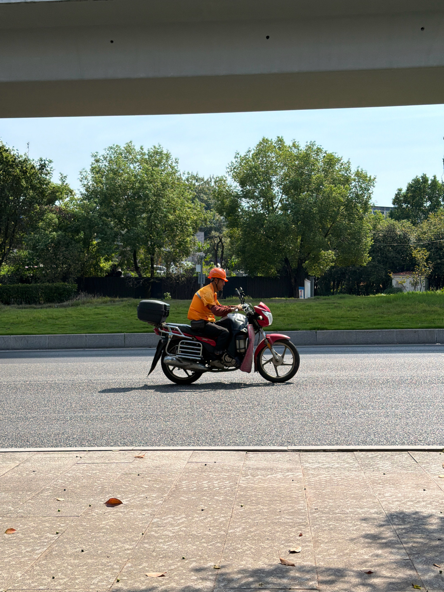 The image depicts a street scene in Jiangjin, China, on a sunny day. A person wearing an orange shirt and an orange helmet is riding a red and white motorcycle on an asphalt road. The motorcycle has a black storage box mounted on the rear. On the left side of the road, there is a grassy area with trees and some bushes. The road is bordered by a concrete sidewalk in the foreground, which is scattered with a few fallen leaves. A concrete structure, possibly a bridge or an overpass, is visible at the top of the frame. The sky is clear and blue.