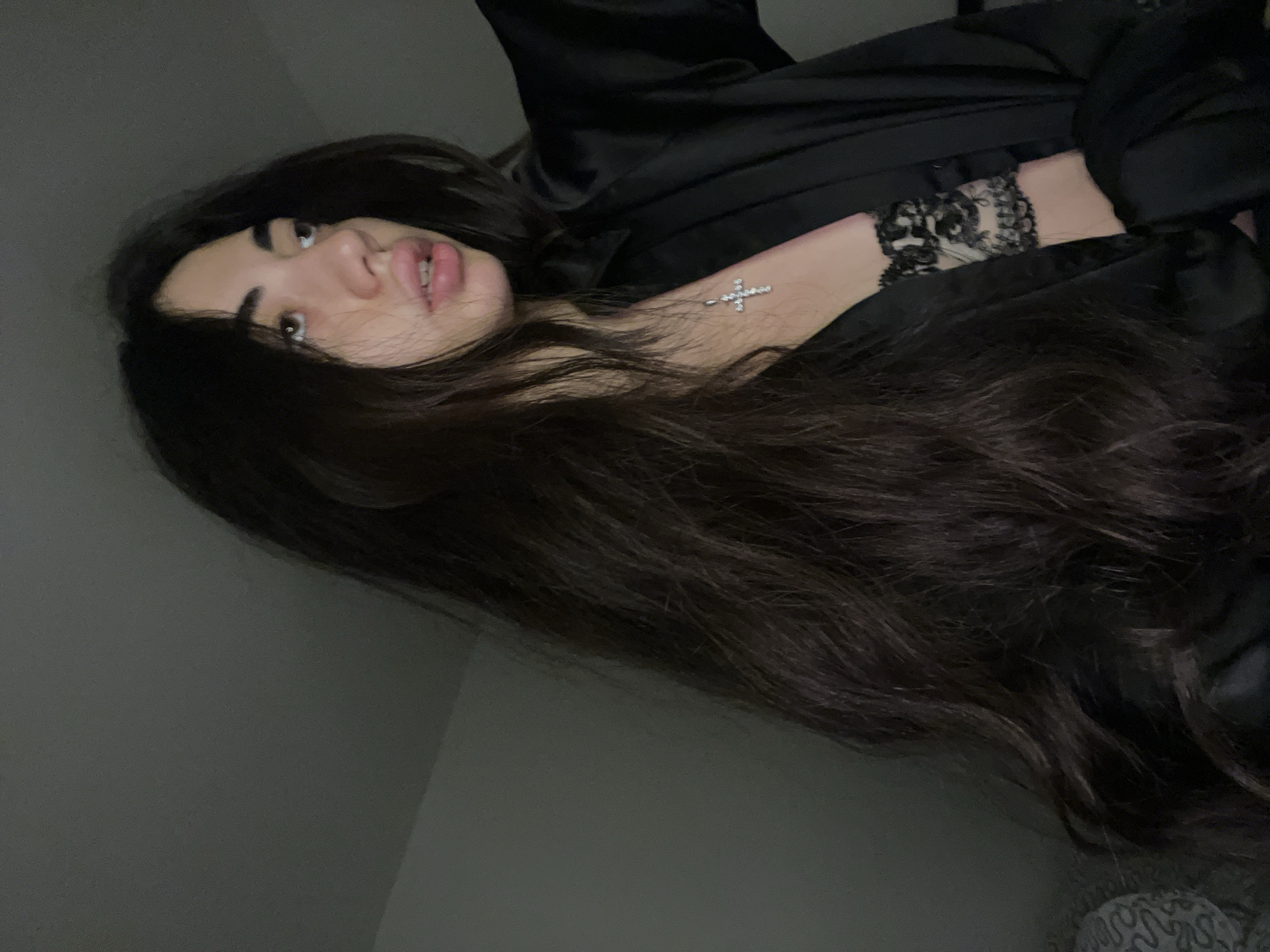 The image shows a young person with long, dark hair, wearing a black satin robe over a black lace bra. They are wearing a silver cross necklace. The person is looking off to the right, with their mouth slightly open. The background is dark and appears to be an indoor setting, possibly a bedroom. There is no information in the image to suggest a location in Chotěšov, Czechia.