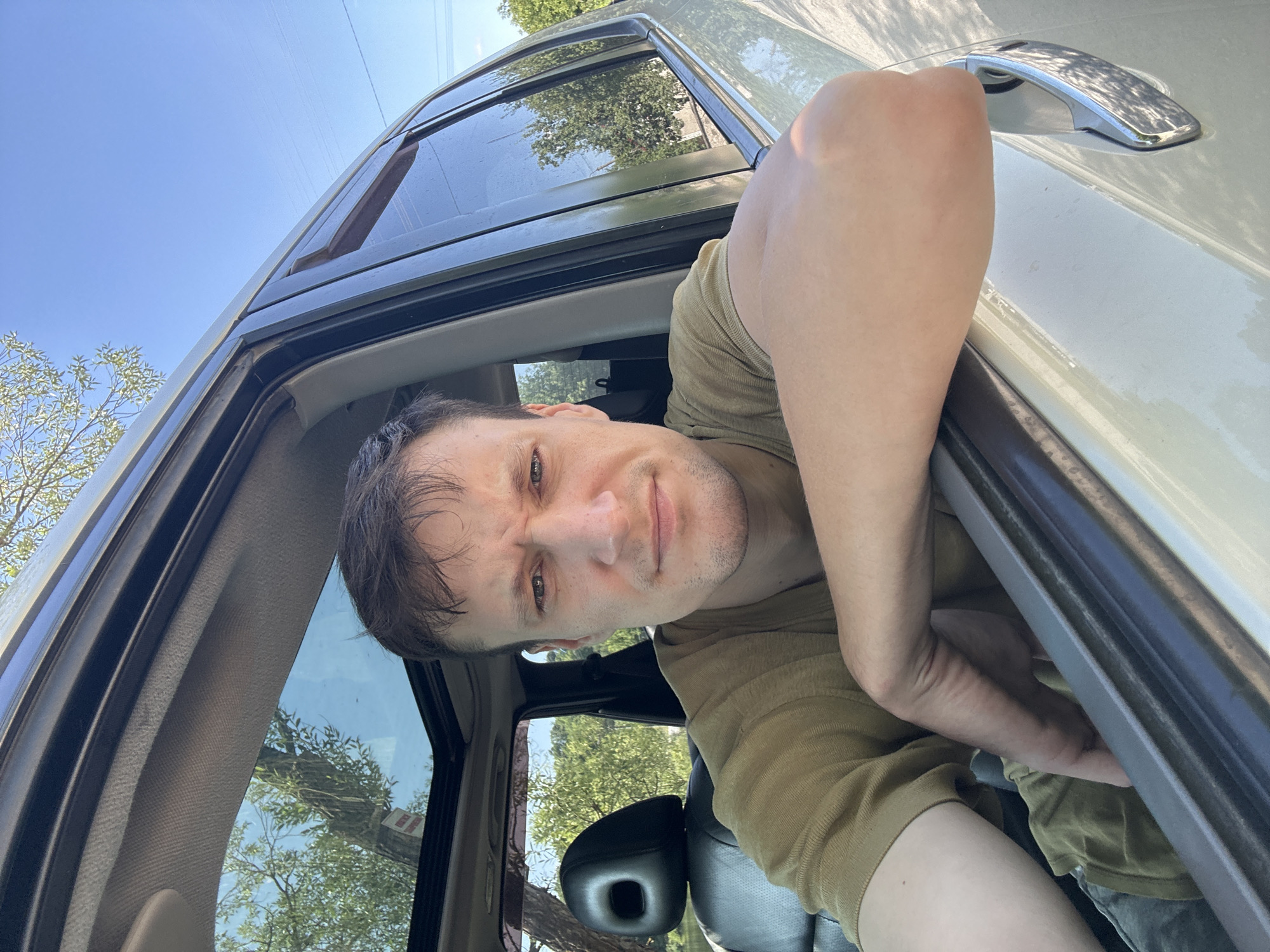 A man is captured in a selfie, leaning out of a car window, his right arm resting on the door frame. He is looking directly at the camera with a neutral expression, his eyebrows slightly furrowed. The interior of the car is visible, including a black leather seat and a sunroof, through which trees and a blue sky are seen. The man is wearing a light olive-colored t-shirt. The car appears to be parked outdoors, with lush green trees and vegetation surrounding it. The presence of power lines in the background suggests an urban or suburban environment. While the specific location within Makiv, Ukraine, cannot be definitively determined from the image alone, the overall scene depicts an ordinary day in a rural or semi-rural setting.