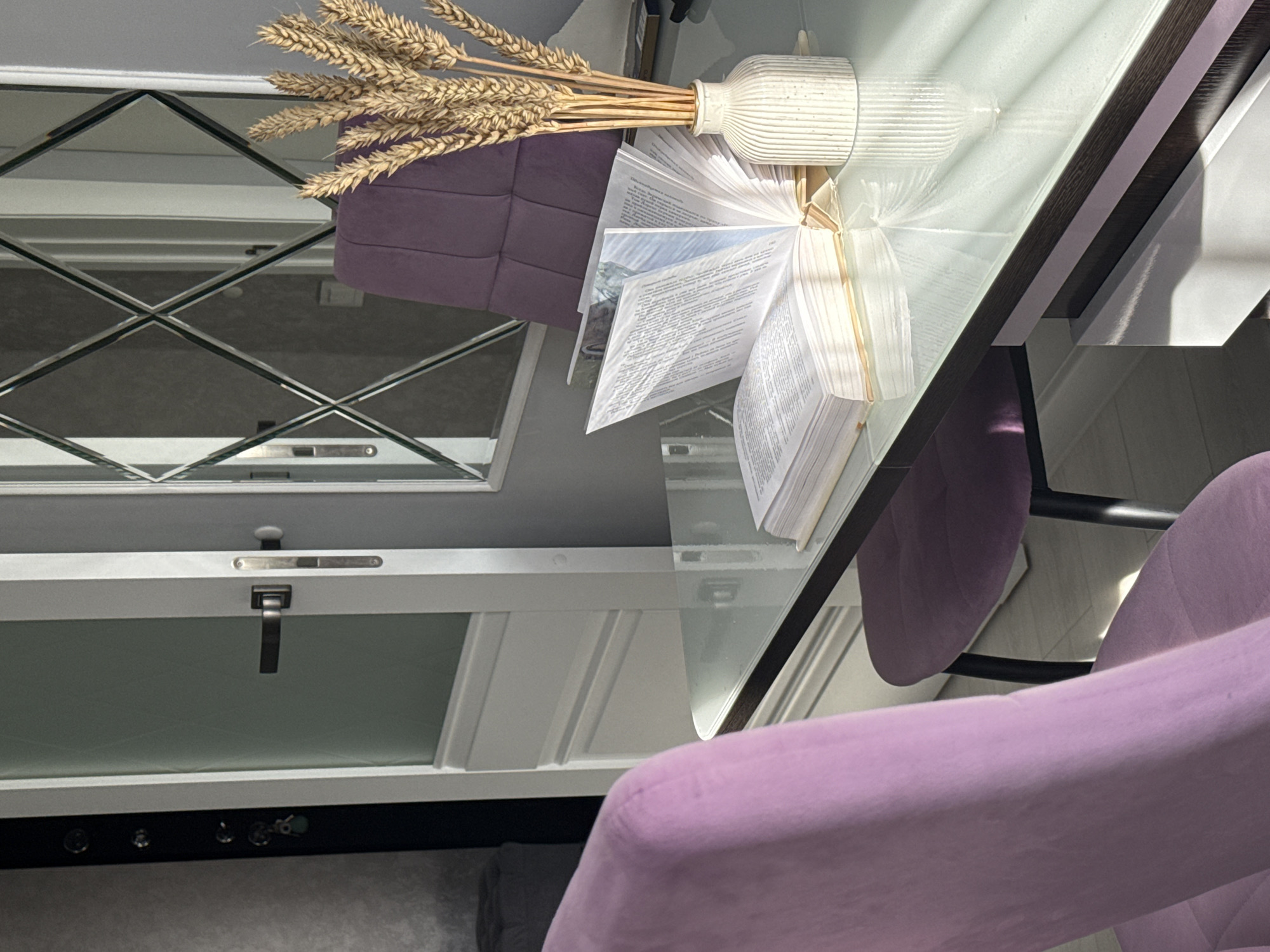The image depicts an indoor scene, likely a home office or study. In the foreground, a light purple chair is visible on the left side. To the right, a glass-top desk is present, on which an open book and a vase with dried wheat stalks are placed. The book is open to pages with text and images, and sunlight streams across the desk and book, creating reflections on the glass surface. In the background, a white door with a glass panel is partially open, revealing a hint of the interior beyond. A large mirror with a diamond-shaped frame hangs on the wall, reflecting the room. Another purple chair is visible in the background, behind the desk. The environment appears to be a well-lit room, with natural light contributing to the overall atmosphere. No people are visible in the scene. The location is described as Unknown, Unknown.