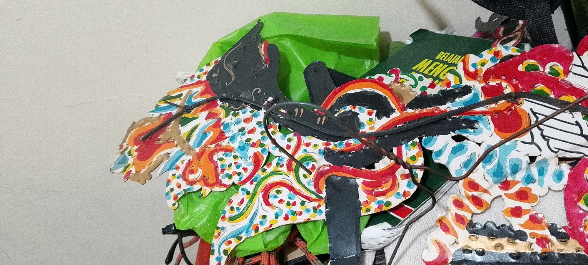 A close-up shot captures a detailed view of a colorful Wayang Golek puppet, likely a traditional Indonesian shadow puppet, against a plain, light-colored wall. The puppet is intricately painted with vibrant colors including red, orange, blue, yellow, and green, featuring a white base with scattered colorful dots. Black accents highlight facial features and decorative elements. The puppet appears to be made of wood or a similar material and is adorned with fabric, possibly green and red. Wires are visible, attached to the puppet, suggesting it's part of a performance or display. In the background, a green object with yellow text, possibly a book or pamphlet with the visible text "BELAJAR MENG", is partially obscured by the puppet. The image is taken indoors in Satriamekar, Indonesia. There are no people visible in the frame.