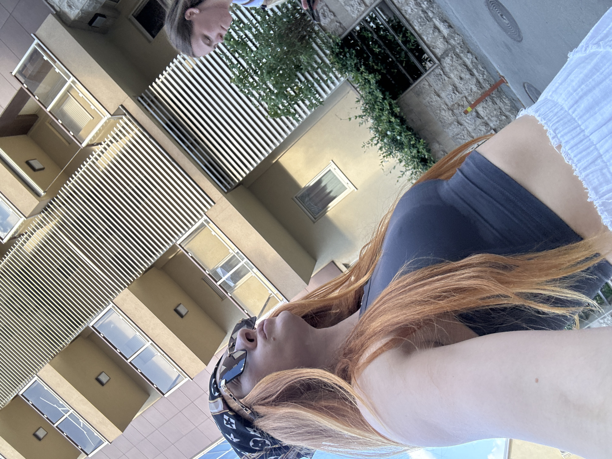 A woman with long, reddish-orange hair, wearing a dark grey tank top, a patterned bandana, and large sunglasses, is positioned in the foreground, looking upwards. Her face is turned to the side, and she has a small mole on her right cheek. To her right and further in the background, a second woman with brown hair is visible from the shoulders up, looking down. They are standing on a paved street with a stone wall and some greenery on the right side. Behind them is a modern building with balconies and horizontal slats. The overall scene suggests an outdoor urban environment, possibly a street in Budva, Montenegro, given the architectural style and the Mediterranean feel of the lighting.