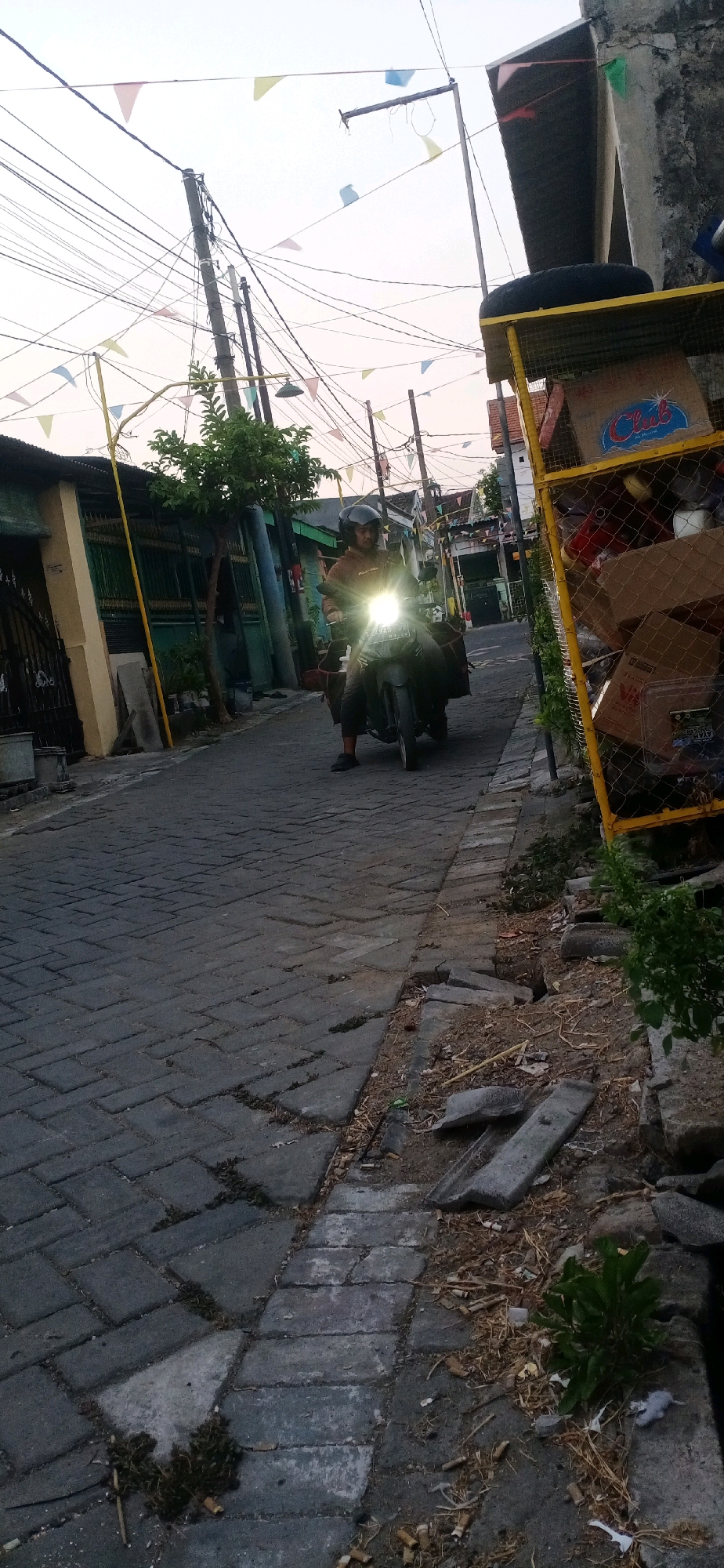 A medium shot captures a person on a motorcycle driving down a narrow, paved street in Sidojangkung, Indonesia. The street is made of dark gray interlocking bricks, and on the right side, there is a yellow metal cage filled with various items, including boxes and what appears to be a tire. The person on the motorcycle is wearing a dark helmet and a light brown jacket. The motorcycle's headlights are on, illuminating the road ahead. Overhead, numerous electrical wires crisscross the sky, and small, colorful triangular flags are strung between poles. The buildings lining the street are visible, with green fences and neutral-colored walls. The overall scene suggests a typical residential street in a town.