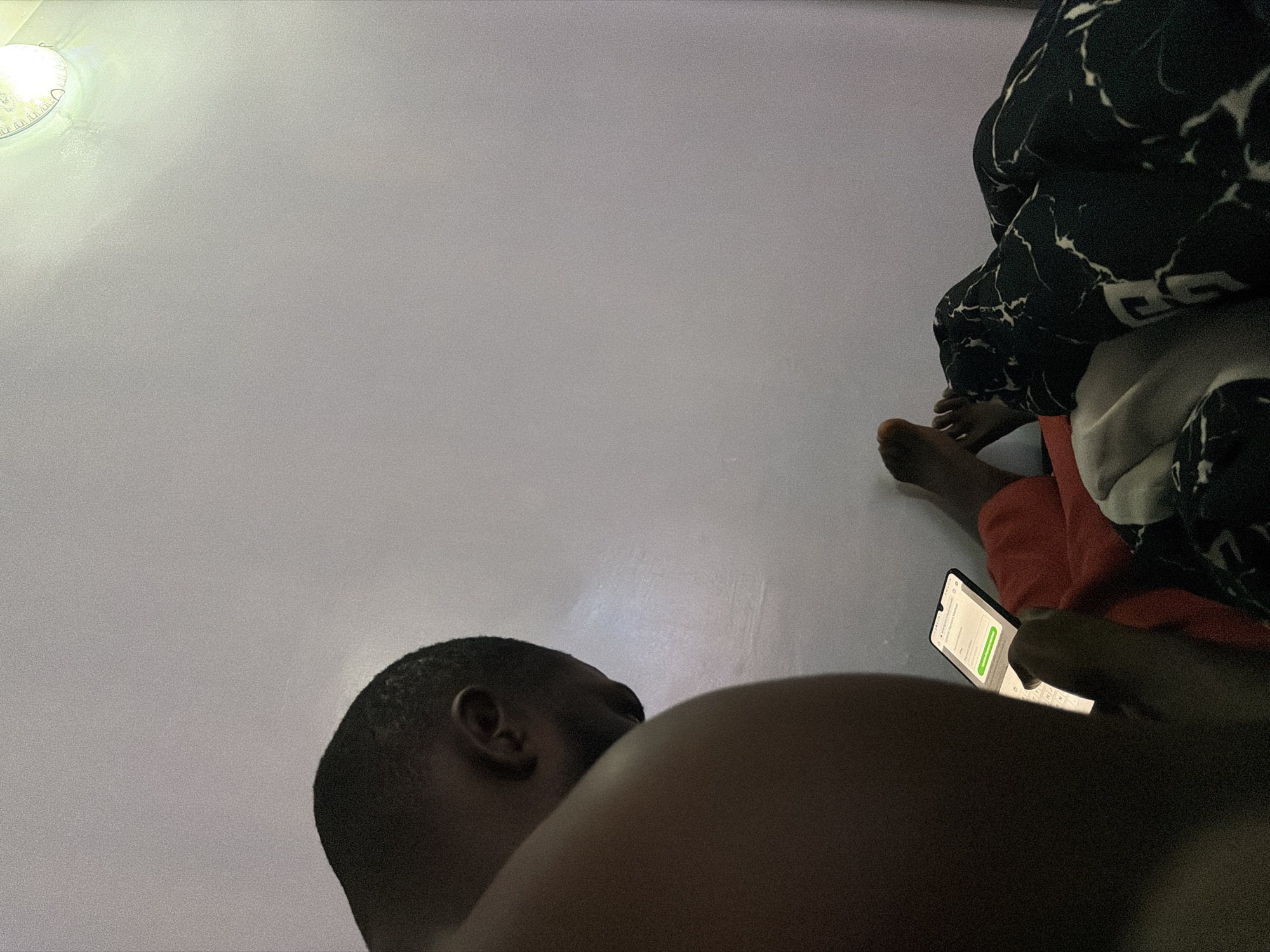 In Barakin Angundi, Nigeria, a dimly lit bedroom scene unfolds. A person, viewed from behind, is seated with their head bowed, engrossed in a smartphone held in their hands. The phone displays a webpage with fields for "Street Address," "City," and "Email," along with a green button labeled "Update Payment Method." Next to them, another person's legs are visible, draped in a dark-colored blanket with a white marble-like pattern. One foot is bare and rests on an orange surface, possibly a blanket or clothing. The background is a plain, light-colored wall with a subtle light fixture visible in the upper right corner, casting a soft glow. The overall atmosphere suggests a private, indoor setting, likely a home.