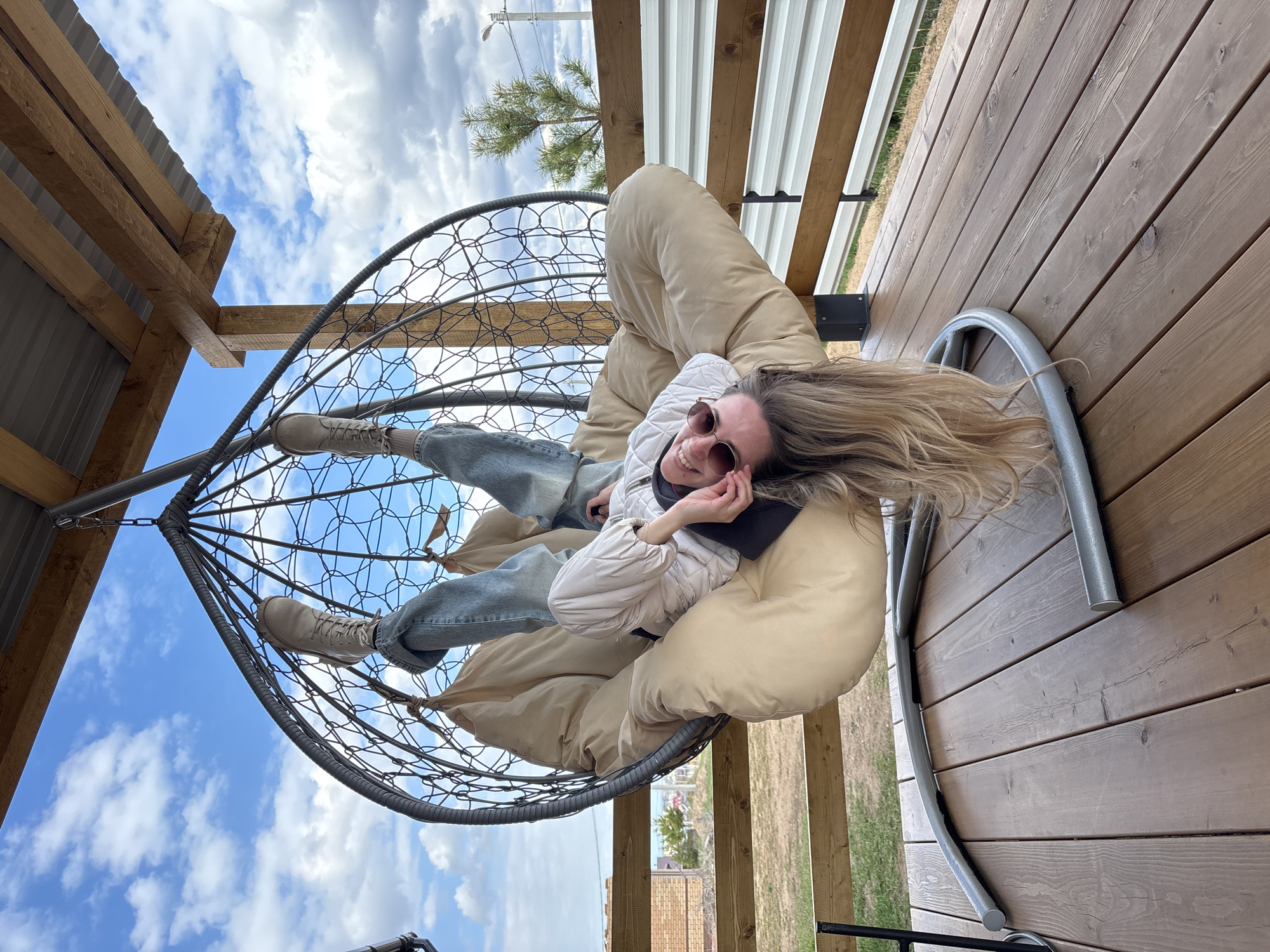 A young woman is depicted relaxing in a modern hanging egg chair on an outdoor elevated wooden deck in Chaikovsky, Russia. She is smiling brightly at the camera, wearing sunglasses, a light-colored puffer jacket, blue jeans, and light-colored lace-up boots. Her long, wavy blonde hair cascades around her head as she reclines comfortably on a large, beige cushion within the chair's woven metal frame.
The setting features a prominent wooden plank deck, along with wooden structural beams and corrugated metal roofing visible overhead, suggesting a sheltered outdoor space. Above and behind the woman, a partly cloudy blue sky dominates the background, indicating daytime with fair weather. Distant elements such as a small pine tree, a utility pole with power lines, and faint outlines of other buildings can be seen, hinting at a semi-rural or recreational area. The woman's relaxed posture and joyful expression convey a sense of contentment. No text is visible in the image.