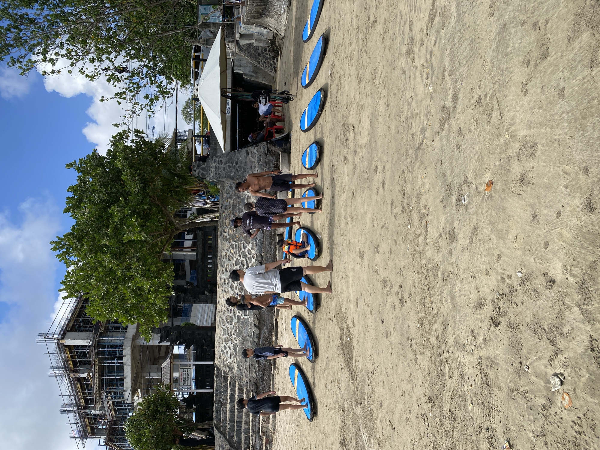In Kuta Selatan, Indonesia, on a sandy beach, a group of people are gathered around several blue surfboards. The group consists of adults and children, some of whom are standing on the boards, while others are standing next to them or sitting. One child is wearing an orange life vest. In the background, a multi-story building under construction is visible, along with trees and a white umbrella. The sky is partly cloudy.