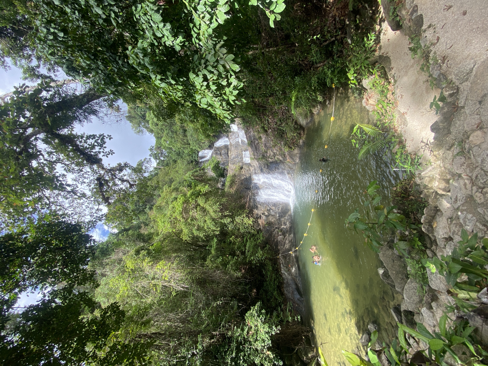 The image depicts a waterfall and pond in Tambon Thai Mueang, Thailand. The waterfall cascades down a rocky slope surrounded by lush green vegetation. A pool of water at the base of the waterfall is clear enough to reveal the pond’s floor. A yellow-buoyed rope stretches across the pool, possibly marking a safe swimming area. Two people are partially submerged in the water. A concrete or stone path edges one side of the pond. Trees and dense foliage dominate the surroundings.