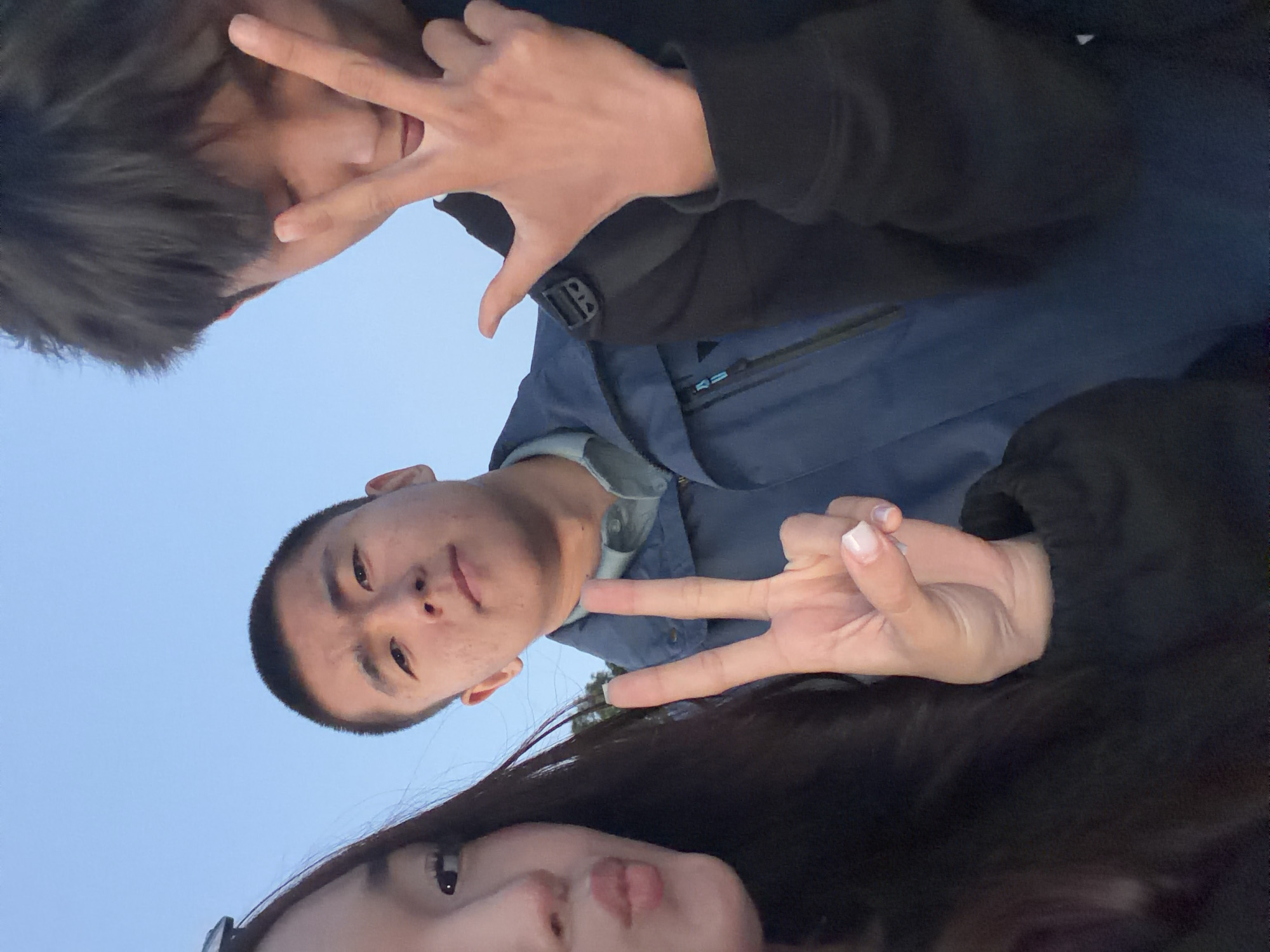 Three young adults are posing for a selfie against a clear blue sky. A woman on the left is pouting her lips, with her left hand visible showing neatly manicured nails and a peace sign. In the center, a man with short hair is looking directly at the camera, wearing a blue jacket over a collared shirt. To the right, another man with dark hair is also looking towards the camera, with his right hand raised in a peace sign. The background is out of focus, showing a hint of greenery and possibly a structure in the distance. The lighting suggests it is either early morning or late afternoon. There is no visible indication of the specific location within Saint Petersburg, Russia.