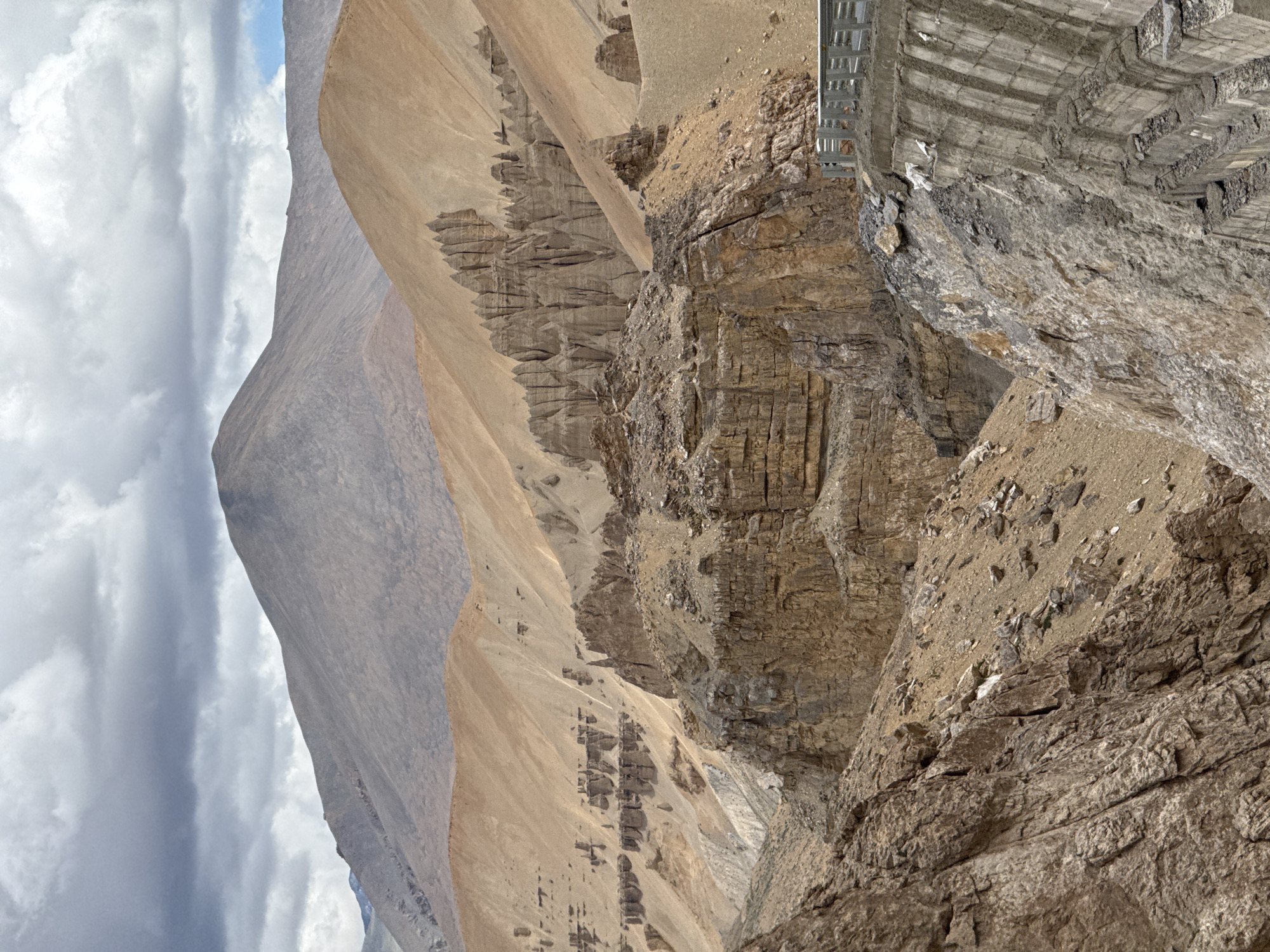 The image depicts a rugged, mountainous landscape in an arid environment. The foreground features a steep, rocky cliff face on the right side, with a concrete structure extending upwards and to the right. This concrete structure appears to be part of a retaining wall or a bridge abutment, with vertical grooving and visible cracks. A metal guardrail runs along the top edge of this structure.

Beyond the immediate foreground, the landscape opens up to reveal a dramatic canyon. The canyon walls are composed of layered rock formations, predominantly in shades of brown, tan, and beige, suggesting a sedimentary origin. There are numerous eroded pillars and spires, indicative of extensive weathering and geological activity. In the middle ground, a prominent ridge displays a series of sharp, vertical rock formations that resemble teeth or natural sculptures.

Further in the background, larger mountains dominate the scene. The closest of these mountains is a large, rounded peak with gentle slopes, appearing to be composed of scree or loose rock. Behind this, a more distant, imposing mountain range rises, its peaks shrouded in a light haze. The sky is overcast with heavy, grey clouds, interspersed with patches of lighter, white clouds, and a hint of blue sky is visible on the far right. The overall impression is one of a vast, remote, and geologically active area. The location is identified as Unknown, India.