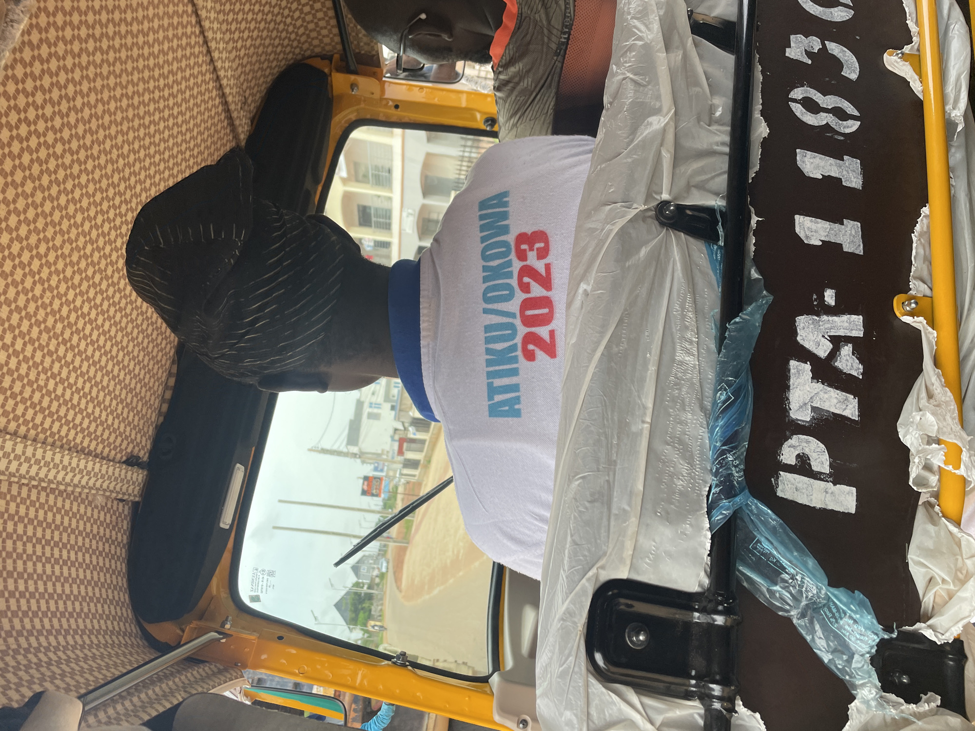 The image shows the interior of a yellow auto-rickshaw, with a person wearing a white t-shirt with the text "ATIKU/OKOWA 2023" printed in blue and red. The person has a black headwrap. Another person wearing glasses and an orange shirt is visible in the background. The auto-rickshaw is in motion, with a road and buildings visible through the windshield. The registration plate of the auto-rickshaw is visible as "PTA-1183G". The setting appears to be Rayfield, Nigeria, as indicated by the context provided. The image is taken from the perspective of a passenger inside the auto-rickshaw.