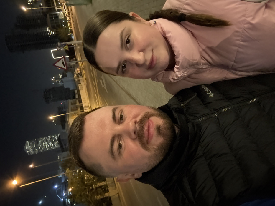 A selfie shows a man and a woman outdoors at night. The man, in a black Columbia jacket, has a beard and short hair. The woman wears a pink puffer jacket, with her dark hair pulled back. Street lights illuminate the scene, casting a warm glow. Tall buildings are visible in the background, suggesting an urban setting. A street sign is also visible behind the woman.