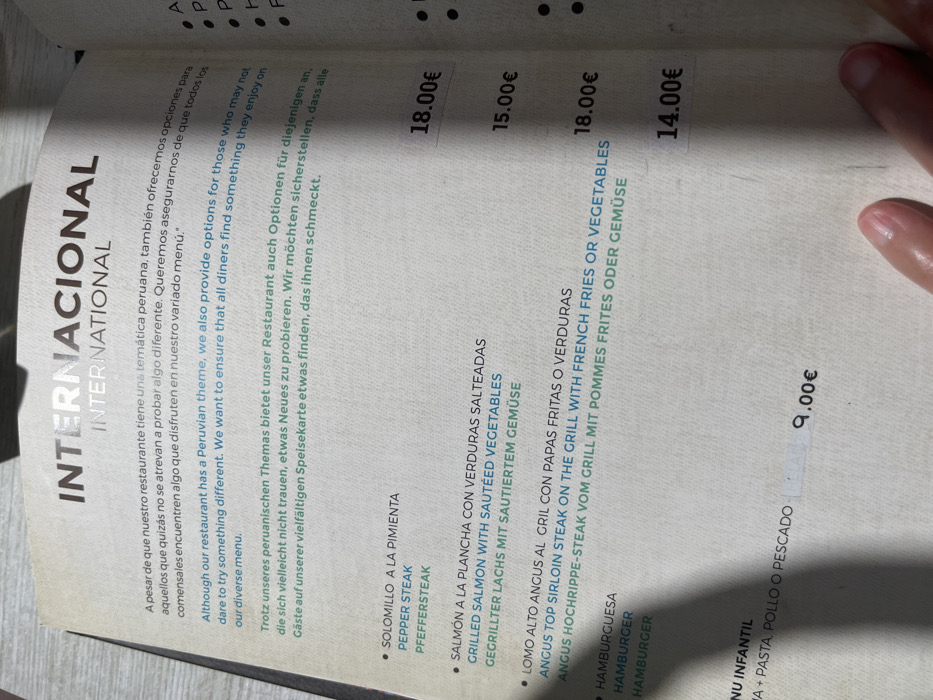 This is a close-up photograph of a restaurant menu, presumably taken by a customer. The menu is printed in black and white with a yellow accent. It is written in Spanish, English and German, and lists a selection of items from an international menu. The menu includes:
- "Solomillo a la Pimienta" (Pepper Steak) for 18.00 Euros
- "Salmon a la Plancha con Verduras Salteadas" (Grilled Salmon with Sauteed Vegetables) for 15.00 Euros
- "Lomo Alto Angus al Grill con Papas Fritas o Verduras" (Angus Top Sirloin Steak on the Grill with French Fries or Vegetables) for 18.00 Euros
- "Hamburguesa" (Hamburger) for 14.00 Euros
- "Menu Infantil" (Kids Menu) for 9.00 Euros.
The menu appears to be on a table with a hand visible in the bottom right-hand corner of the image. The image was taken in a restaurant in Las Palmas de Gran Canaria, Spain, at the coordinates 28.147, -15.43.