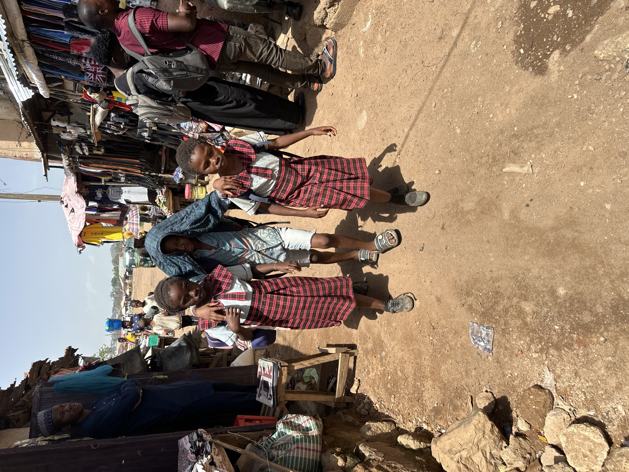 A group of people, including children, are gathered in what appears to be an outdoor market in Bukuru, Nigeria. In the foreground, three children are visible. One child, wearing a red and white plaid dress and a light-colored shirt with a collar, stands with their arm around another child. The second child, also wearing a similar red and white plaid dress, is positioned behind the first. A third child, wearing a patterned shirt and shorts, stands next to them. The children are standing on a dirt ground, with some scattered rocks and debris.
Behind the children, there is a stall with clothing items hanging on display, suggesting a market setting. The stall has a corrugated metal roof. Further in the background, more people and market stalls are visible, along with some vehicles. The scene is illuminated by bright sunlight, indicating it is daytime, likely mid-morning or afternoon, with clear weather. The overall impression is a bustling market scene, possibly during a regular market day. The expressions on the faces of the children are neutral or slightly focused, not conveying strong emotions. No visible text is clearly discernible in the image.