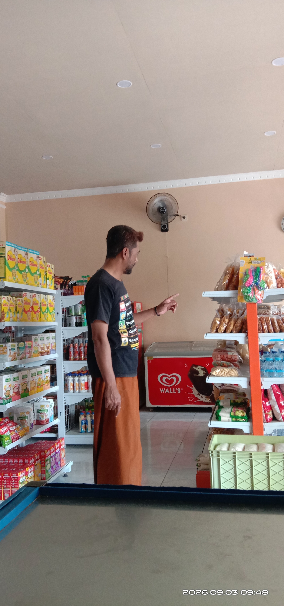 In a retail store in Pakong, Indonesia, a man wearing a dark t-shirt and an orange sarong stands facing away from the camera, looking to his right. He is gesturing with his right hand, his index finger extended. In the background, shelves are stocked with various products, including packaged goods like milk powder and snacks, as well as bottled drinks. An ice cream freezer with the "Wall's" logo is visible to the right of the man. The environment appears to be well-lit, with ceiling lights and a wall-mounted fan. The timestamp "2026.09.03 09:48" is visible in the bottom right corner of the image.