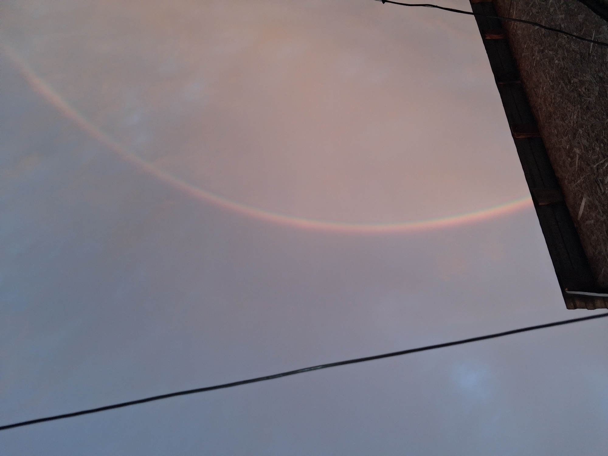 This image depicts a rainbow arcing across a cloudy sky. In the foreground, the corner of a structure, possibly a building with a corrugated roof, is visible on the left side of the frame. A dark horizontal wire stretches across the upper portion of the image. The sky exhibits soft hues of pink, purple, and blue, suggesting either dawn or dusk. The scene appears to be an outdoor environment, likely in a semi-urban or rural setting given the presence of the building and wires. The image was taken in Semey, Kazakhstan.