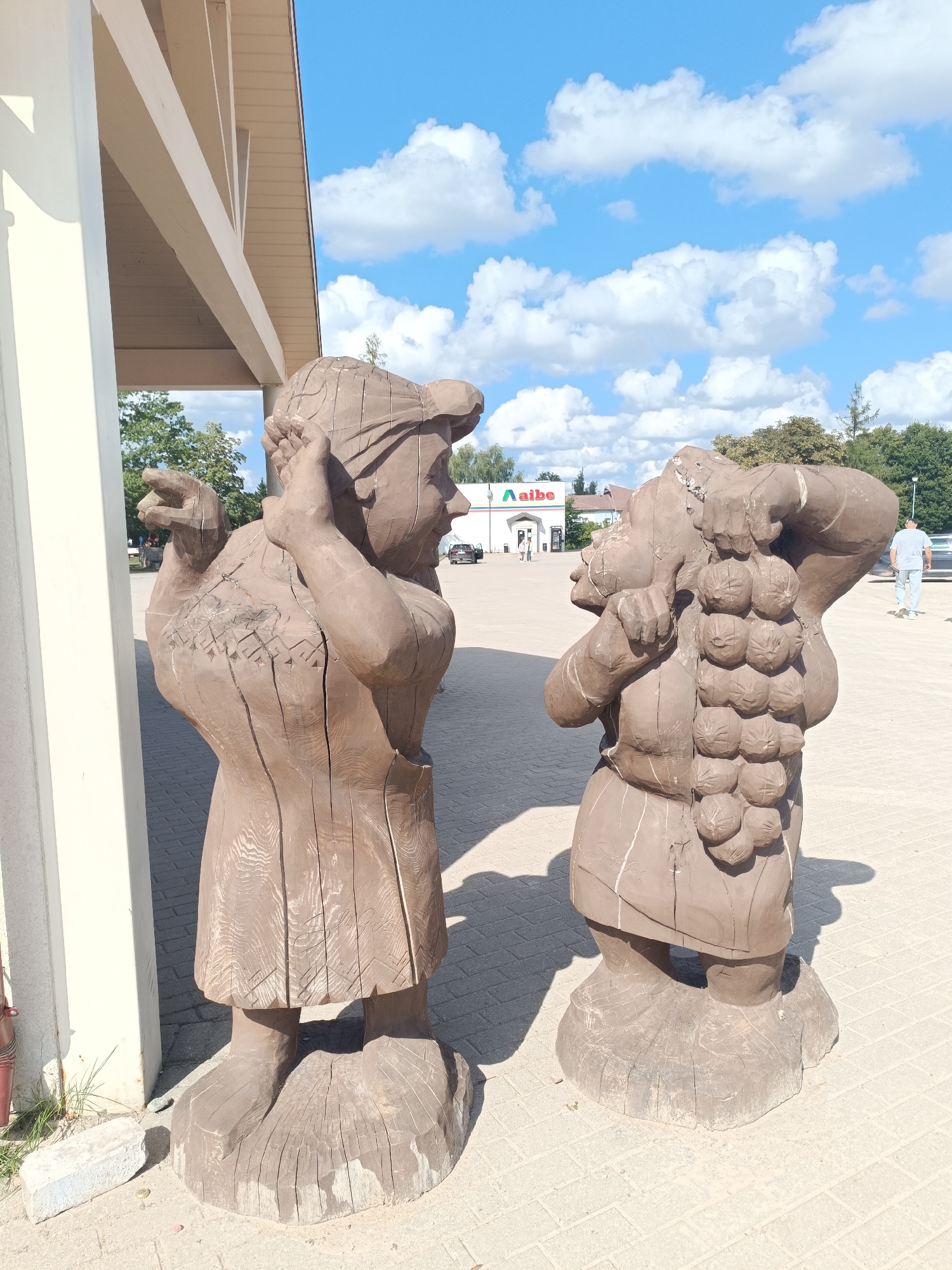 Two wooden sculptures of figures are positioned in front of a building with a sign that reads "Aibe" in Aizpute, Latvia. The sculpture on the left depicts a person wearing a headscarf and a dress, with their hands raised to their head and a finger extended forward. The sculpture on the right appears to be carrying a cluster of grapes on its shoulder. The figures are carved from wood and stand on rounded bases. They are located on a paved area, possibly a sidewalk or plaza, in front of a store. In the background, trees and a blue sky with white clouds are visible. A few people are present in the background, and a car is parked in front of the store.