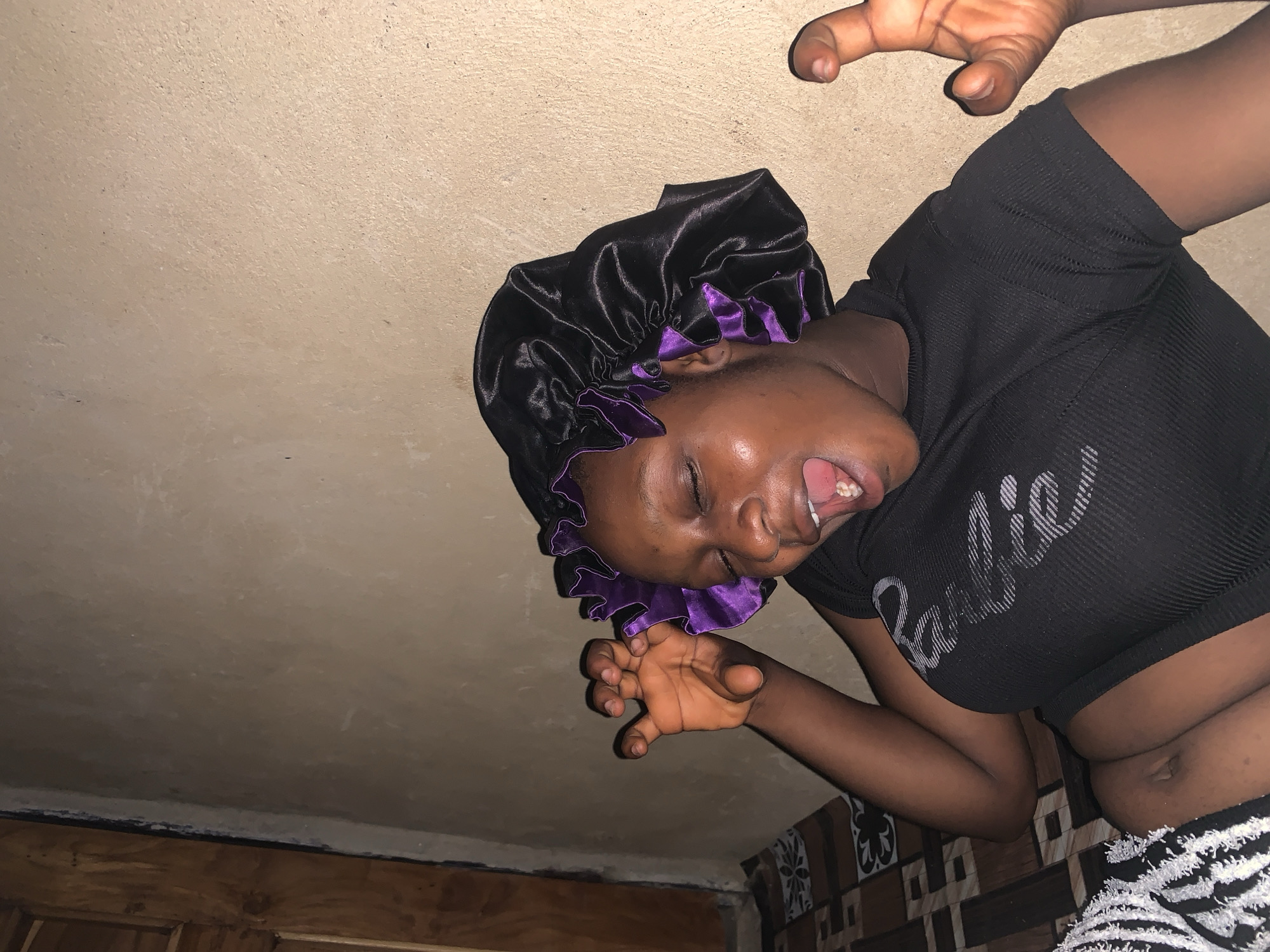 In a dimly lit indoor scene, a young Black woman is lying down, her body angled slightly towards the viewer. She is wearing a black satin bonnet with purple trim, and a black ribbed t-shirt with white lettering that reads "Barbie." Her eyes are closed, her mouth is open in a wide yawn or shout, and her tongue is visible. Her right arm is raised, with fingers spread in a claw-like gesture. Her left arm is bent, with her hand near her head. The background consists of a textured, light-colored wall and a wooden door or panel on the left. The floor beneath her is covered with a patterned mat or rug. The overall impression is one of playful exuberance or exaggerated expression.