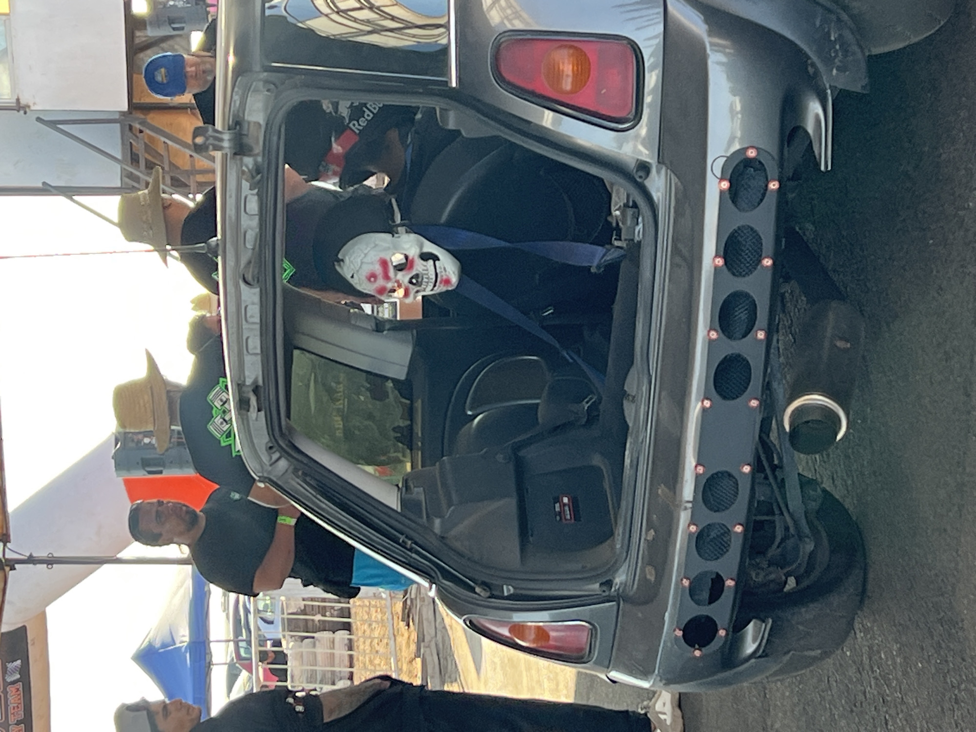 The image shows the open hatchback of a gray car. A white skull mask rests on the backseat, secured by the seatbelt. The car has a modified rear bumper featuring a black panel with circular cutouts. A chrome exhaust pipe is visible near the rear tire. Several people stand around the car, including a person in a black shirt with arms crossed. In the background, there are event tents and metal scaffolding. This event likely took place outdoors, potentially in Arica, Chile, based on the request.