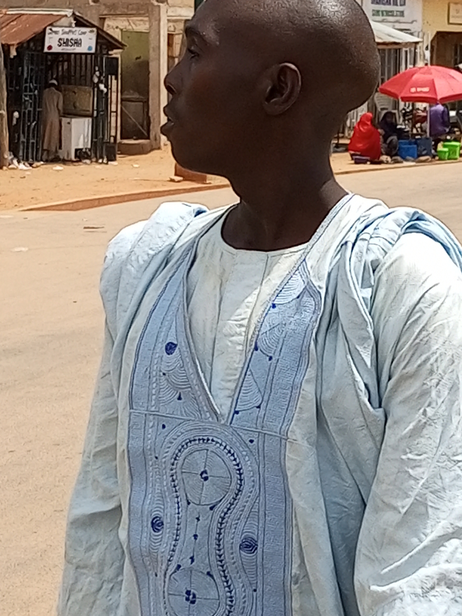 In Gombe, Nigeria, a man in a light blue garment with intricate blue embroidery stands on a dusty street. He is seen from the shoulder up, with his head shaved and turned to the right. In the background, a small stall with a sign that reads "SHISHA" is visible, with a person in a long brown robe standing inside. Further in the background, a red umbrella shades a group of people, and more stalls and buildings line the street. The overall scene depicts a vibrant street market environment in the city.