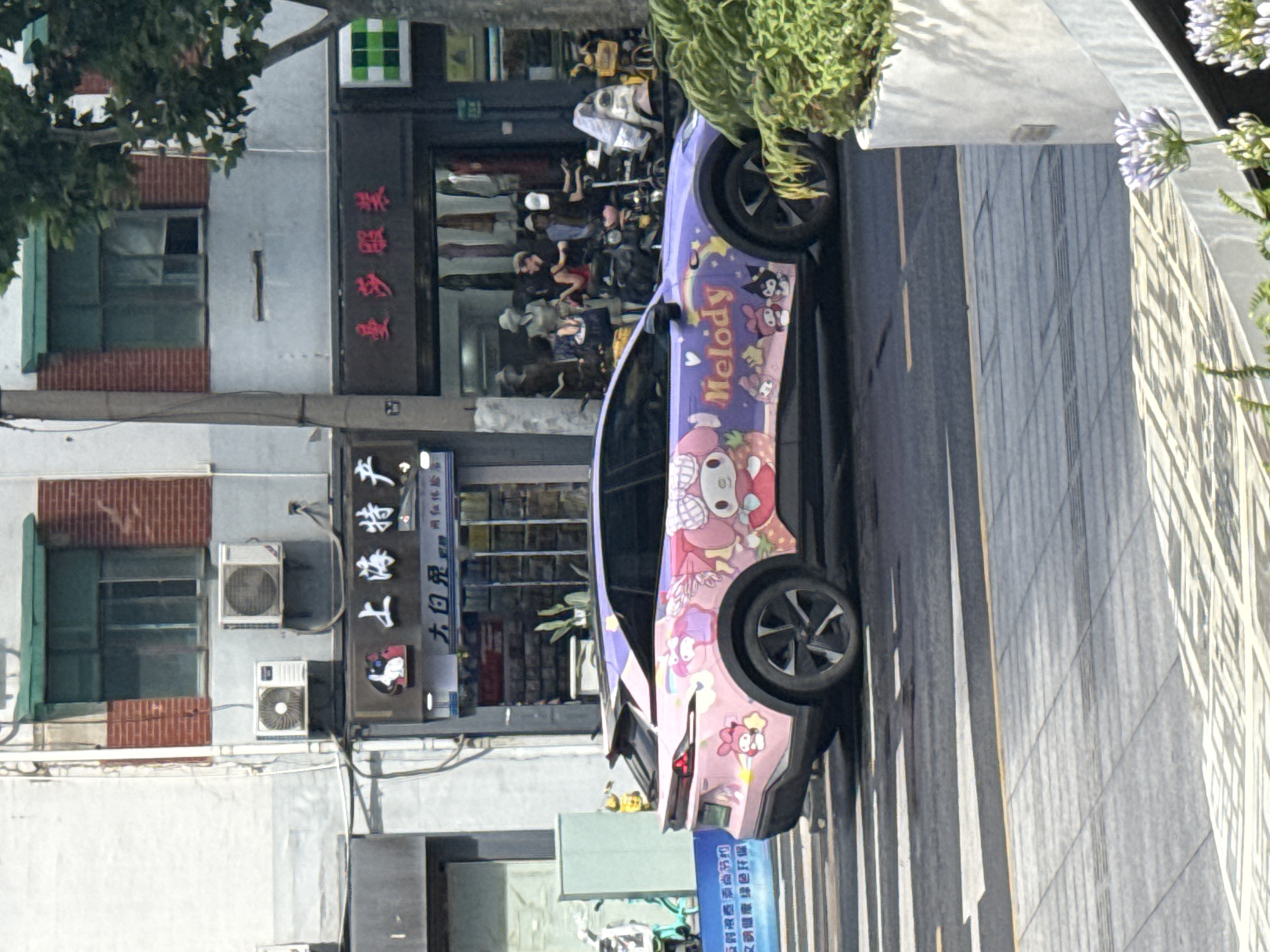 An exterior, eye-level, medium shot captures a car wrapped with a pink and purple design featuring cartoon characters from the Sanrio franchise, specifically My Melody, driving on a street in Shanghai, China. The car is a modern SUV with a black roof and black rims on its wheels. The street is paved with asphalt, marked with white pedestrian crossing lines and a yellow line along the curb. To the left of the car, a blue banner with Chinese text is partially visible, attached to a building. The building behind the car has a gray facade with red brick accents and green awnings over windows. There are air conditioning units mounted on the building's exterior. The ground level of the building features storefronts with clothing displays and mannequins visible through the windows. To the right of the car, a white planter box contains lush green foliage and purple flowers. A tree with green leaves is also present on the right side of the image, partially obscuring the building. The overall scene appears to be a daytime shot on a city street.