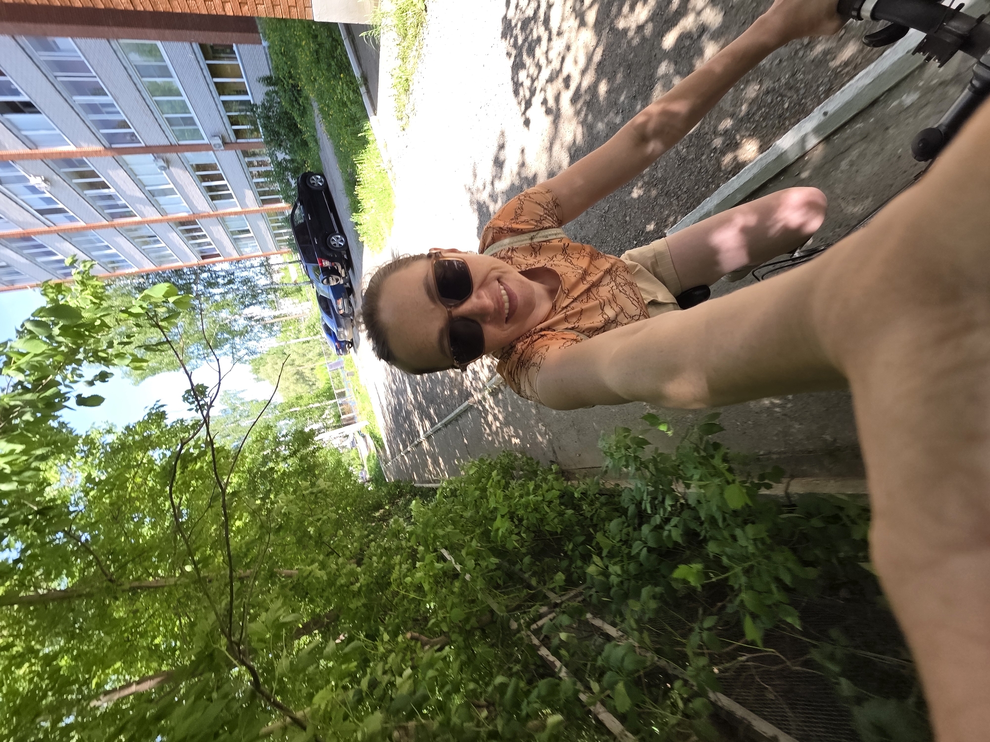 A woman wearing sunglasses and an orange shirt takes a selfie while riding a bicycle on a path next to a building with many windows. Two cars are parked near the building. Trees line the path. The location may be Chaikovsky, Russia.