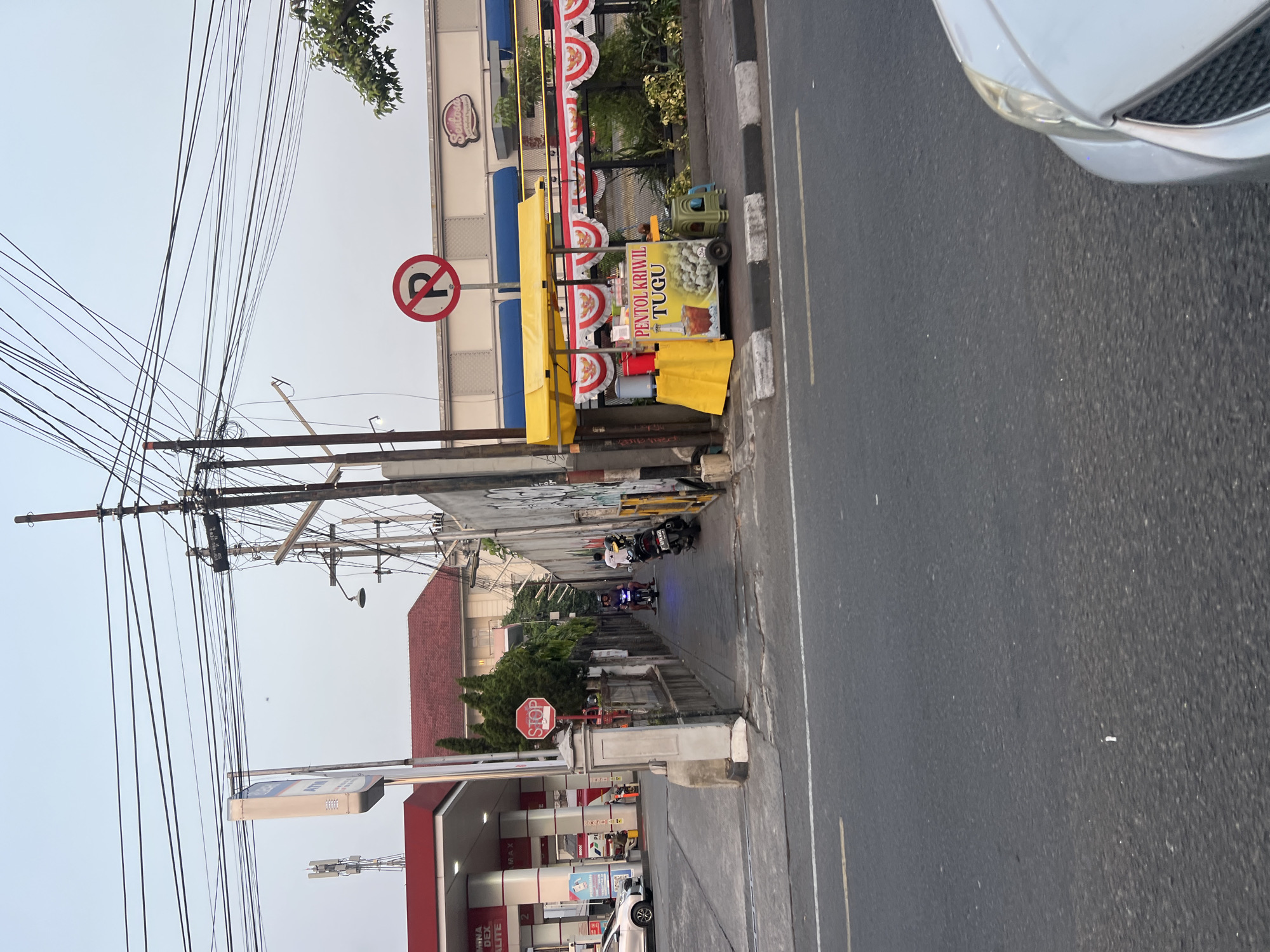 The image captures a street scene in Yogyakarta, Indonesia, likely during the late afternoon or early evening, given the lighting. On the left side of the frame, a Pertamina gas station with its signature red canopy is visible, featuring a prominent ATM sign. To the right of the gas station, a narrow sidewalk runs alongside a wall adorned with graffiti. Several motorcycles are parked or being ridden along this sidewalk, with two individuals on one motorcycle. A STOP sign is mounted on a pillar near the entrance to the sidewalk.
Further down the street, on the right side, a yellow food cart with a red and white decorative banner is set up. The cart displays text indicating it sells "PENTOL KRIWIL TUGU," a local snack. A red and white "no parking" sign is also visible on a pole near the food cart. In the background, a building with a red roof and a sign that reads "Sentosa" can be seen. The street itself is paved asphalt with visible lane markings. The foreground shows the front right portion of a silver car, suggesting the photo was taken from another vehicle. The sky above is overcast, and numerous overhead power lines crisscross the scene, supported by utility poles.