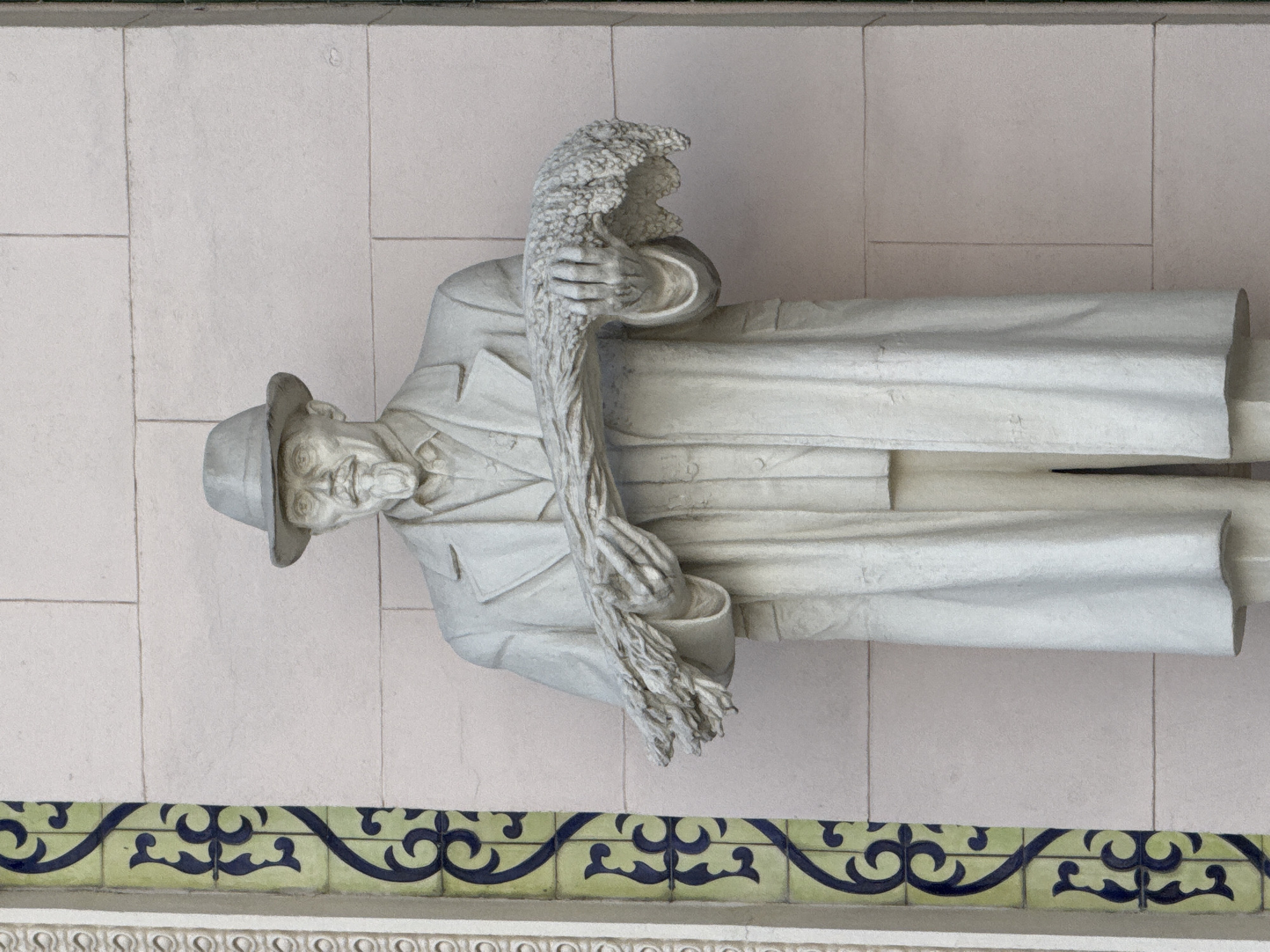 This is a full-shot, eye-level, outdoor, daytime photograph of a white stone statue of a man standing in front of a light-colored brick wall. The man is wearing a hat, a coat, and a vest. He has a mustache and a goatee and is holding a bundle of wheat in his arms. The statue is located in front of a wall adorned with decorative green and blue tiles. The overall scene is calm and depicts a solitary figure in a public space.