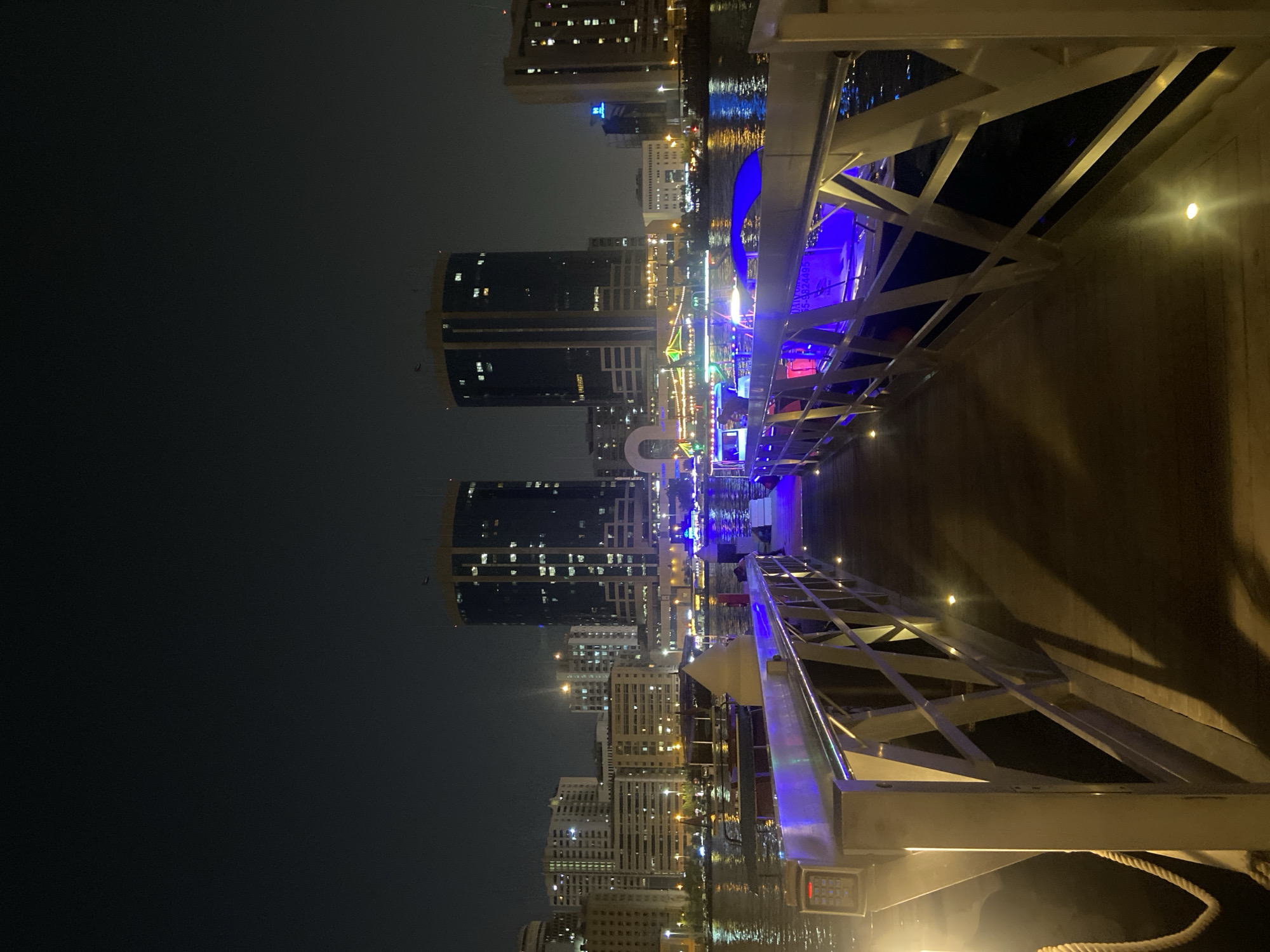 A night view captures a waterfront scene in Deira, United Arab Emirates, likely near the Deira Creek. The foreground features a wooden pier with metallic railings, illuminated by soft yellow lights embedded in the walkway. A thick rope is visible on the left side of the pier. Beyond the pier, a body of water reflects the lights of the cityscape. Several boats are docked or cruising on the water, some adorned with colorful lights, particularly blue and purple, suggesting a nighttime cruise or entertainment venue. In the background, towering modern buildings with illuminated windows punctuate the dark night sky, indicative of a bustling urban environment. The architecture of the buildings suggests a contemporary city skyline.