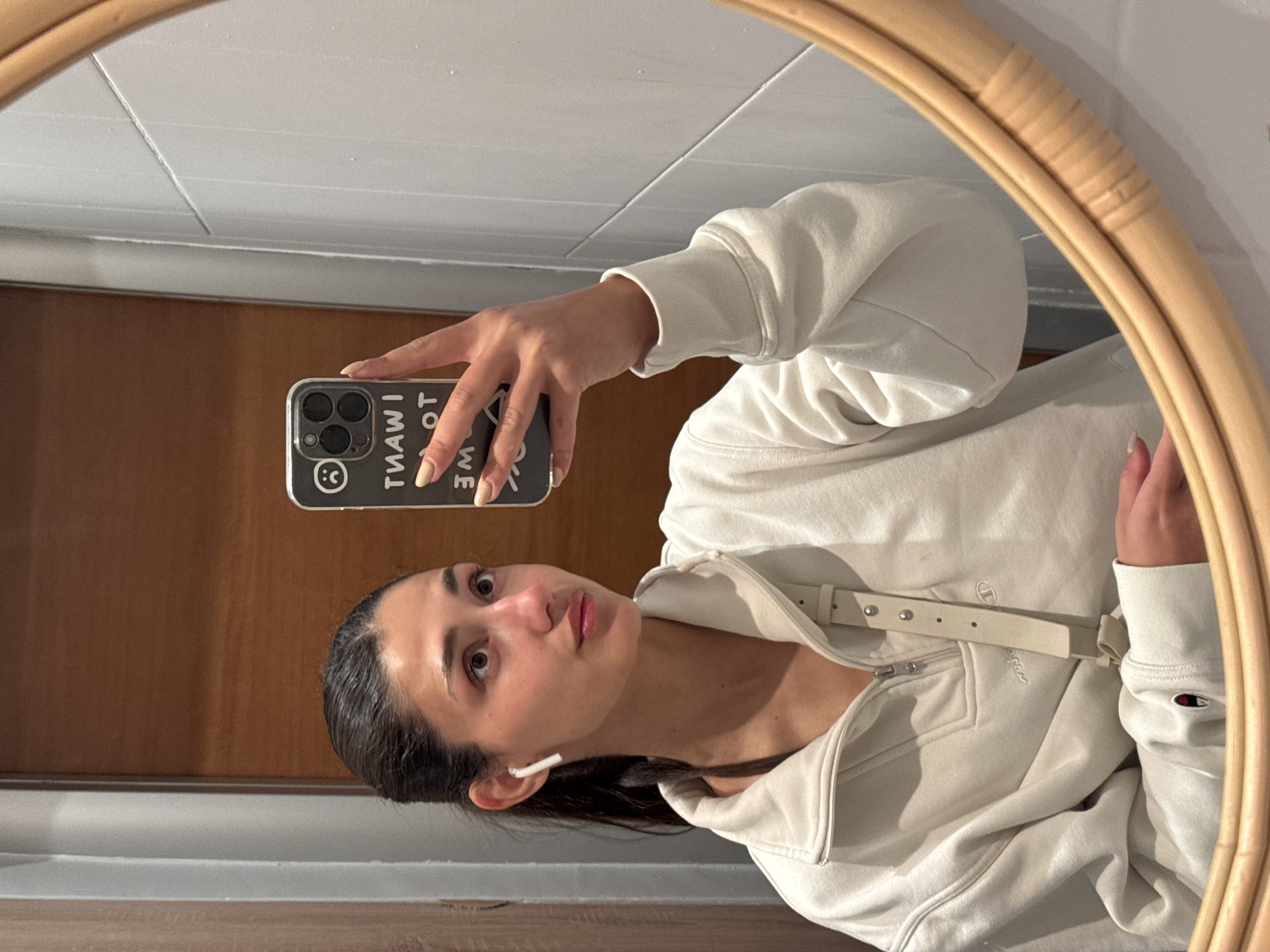 A young woman stands in front of a mirror in a bathroom-like setting with a white-tiled wall above a wooden panel. The mirror has a light-brown woven frame. She is wearing a light-colored, zip-up hoodie with a belt and holds a phone with a black case that reads "I WANT TO WE". She is looking towards the mirror and holding the phone up to take a selfie. An earbud is visible in her ear. The location cannot be confirmed as being in Bar, Montenegro, based on the visual data.