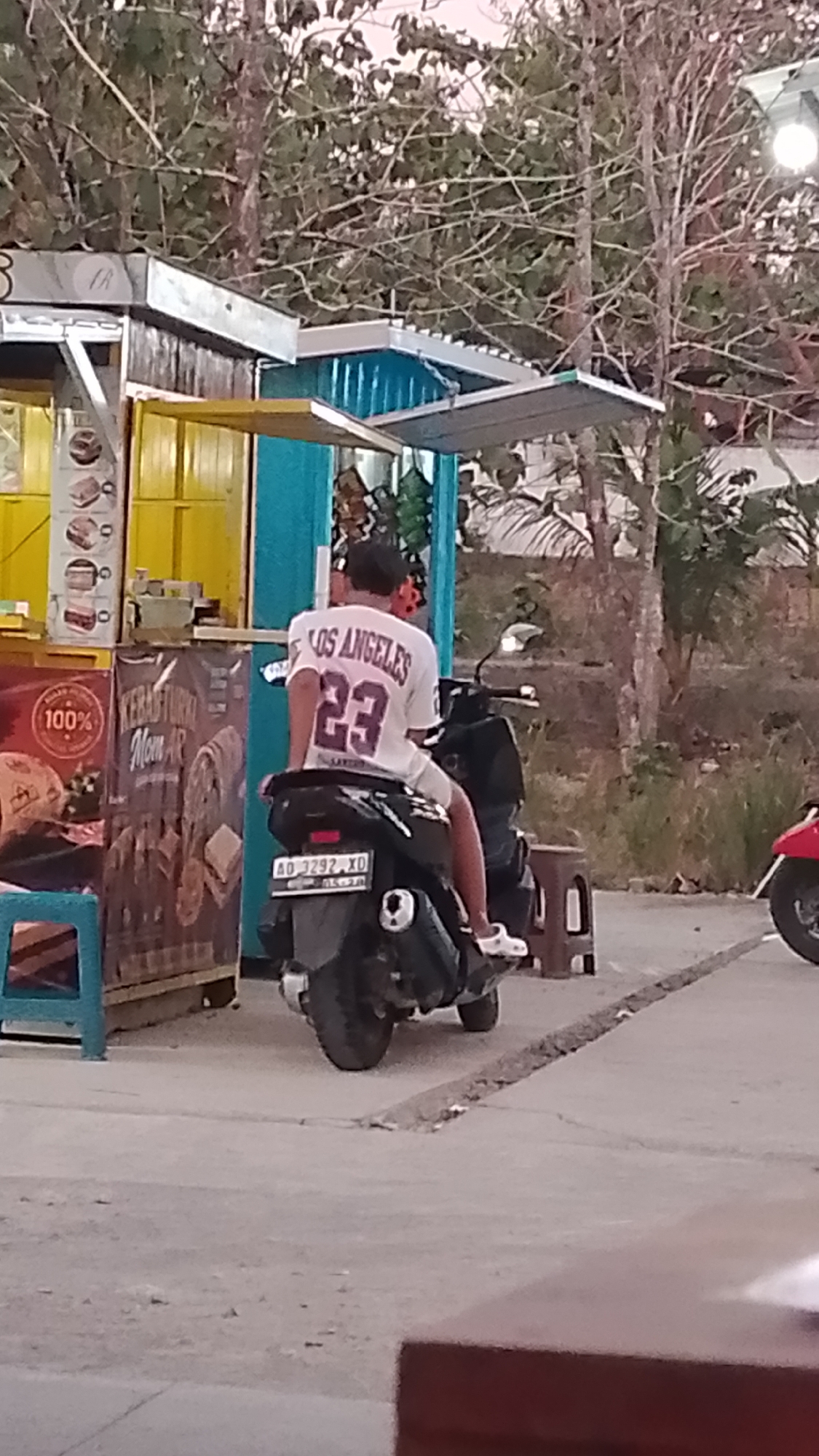 A full shot captures a man sitting on a black motorcycle, facing away from the camera. He is wearing a white t-shirt with "LOS ANGELES 23" printed on the back in purple. He is also wearing white shorts and white slip-on shoes. The motorcycle has a license plate that reads "AD 3292 XD".
In the background, a food stall is visible. It has a yellow front with blue accents and a metal roof. Various items are displayed in the stall, including what appear to be packaged snacks or food items. A small blue stool is placed to the left of the stall.
The ground is paved with a light-colored concrete. Trees with sparse foliage and branches are visible behind the food stall. The overall lighting suggests it might be late afternoon or early evening. The image appears to be taken in Karanglangu, Indonesia, given the license plate's format.