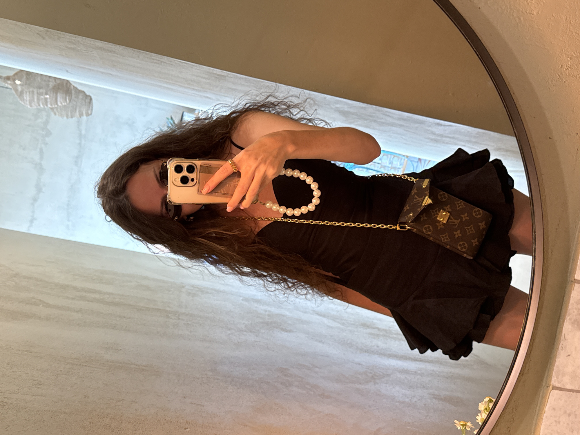 A woman with long, curly brown hair and dark sunglasses is taking a mirror selfie. She is wearing a black dress with a ruffled hem and a pearl necklace. She is also holding a smartphone in her right hand and has a small designer crossbody bag with a gold chain and LV monogram print slung over her shoulder. The background appears to be an indoor setting with textured walls and a large oval mirror. The location is not identifiable as Moscow, Russia.