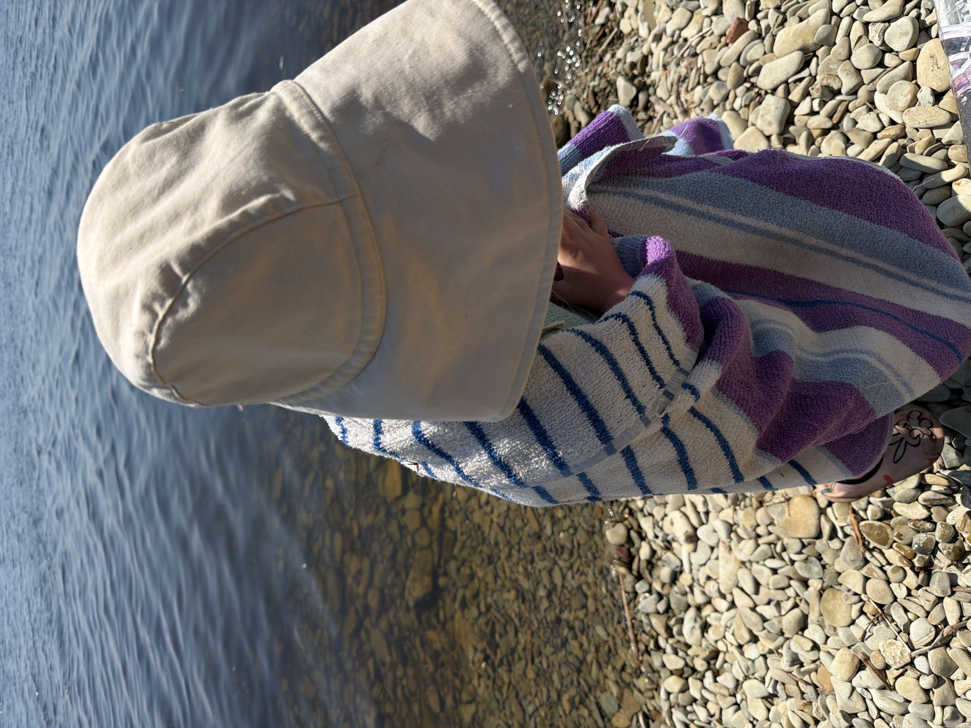 A child, wearing a wide-brimmed sun hat and wrapped in a striped towel, sits on a rocky shoreline, looking out at the water. The water is calm and reflects the sunlight. The shoreline is covered in smooth, light-colored pebbles. The child's feet, wearing pink sandals with a floral design, are visible. The scene is set on the banks of the Volga River in Kamyshin, Russia, under a clear, sunny sky.