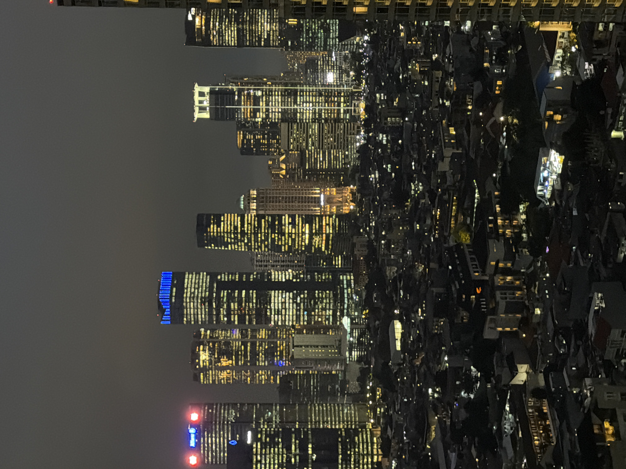 The image depicts a nighttime view of South Jakarta, Indonesia, featuring a stark contrast between high-rise buildings and densely packed residential areas. The city's skyline is punctuated by illuminated skyscrapers, with numerous windows creating a grid-like pattern. Some buildings display blue and red decorative lighting. Adjacent to the skyscrapers, a lower-rise residential area is visible, characterized by clustered buildings with sporadic lighting. The overall scene is a mix of modern urban development and traditional neighborhood structures, illuminated against a dark sky.