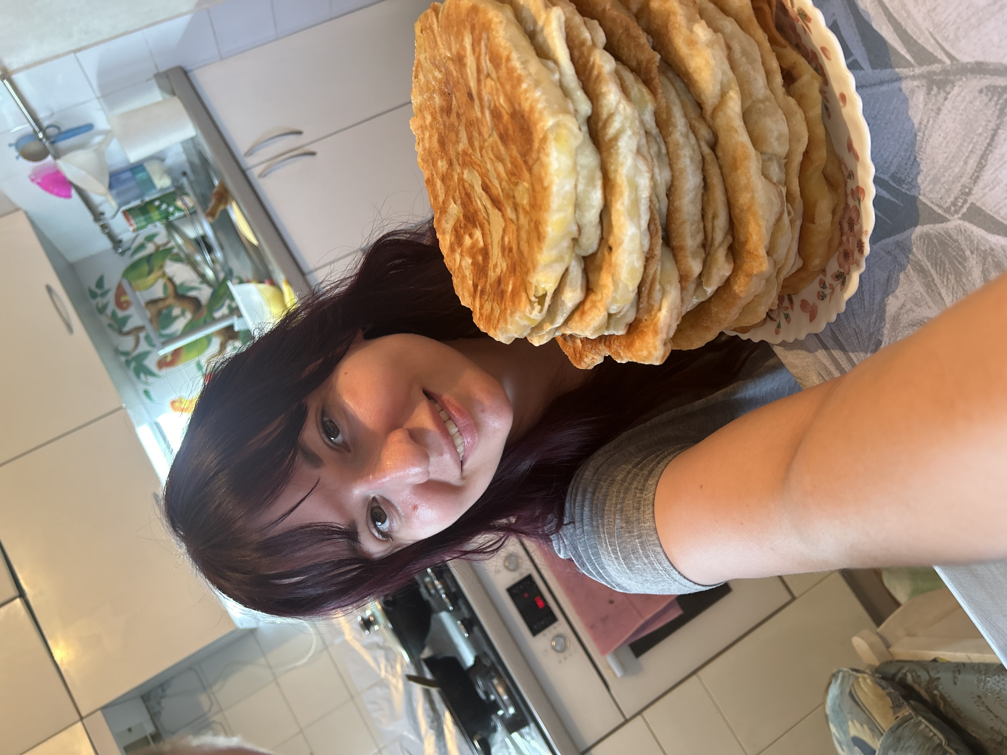 A Caucasian female, appearing to be in her 20s or 30s, with dark reddish-brown hair and bangs, is depicted smiling directly into the camera in a selfie. She wears a grey, ribbed short-sleeved garment. Her right arm extends forward, supporting a white plate with a pink floral design, which holds a vertical stack of approximately ten to twelve round, pan-fried flatbreads with a golden-brown, textured surface. The scene is set in a kitchen located in Фоки, Russia. The background features white tiled walls and a light-colored countertop. Overhead and base cabinets are white with silver bar handles. To the left, a white electric range and oven unit is visible, displaying "080" on its digital interface. Two black cooking pans are positioned on the stovetop, and a pink fabric towel is suspended from the oven door handle. Above the sink area, a decorative tiled border with a motif of green parrots perched on tree branches with orange elements is present. A metal rail system is affixed to the wall, supporting various kitchen utensils, including a white plastic funnel, a pink cleaning tool, and a blue-handled item. The lighting in the kitchen is bright and appears to be evenly distributed.