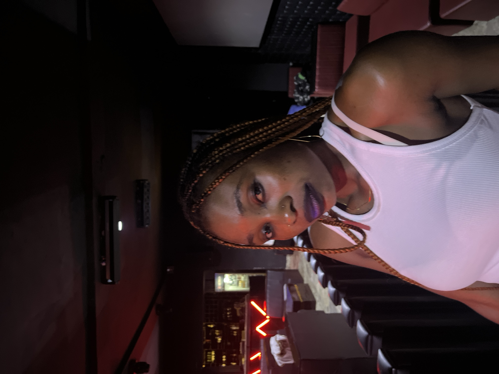In Howayi, Nigeria, a woman with braided hair and dark lipstick poses for a photo indoors. She is wearing a white tank top. The background features dark seating, possibly in a cinema or lounge, with red accents and lighting. A bar area with bottles is visible to the left.