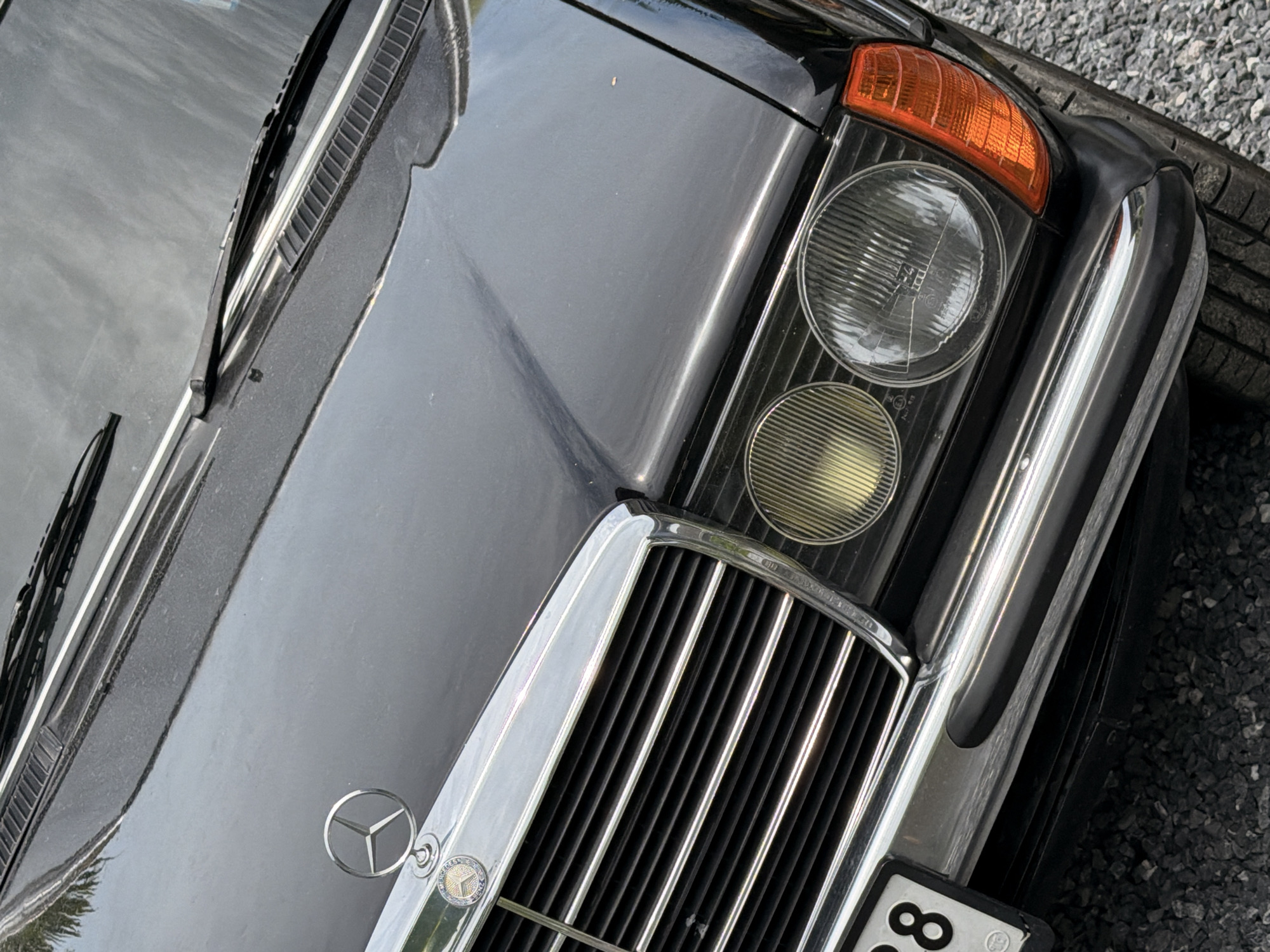 The image shows the front of a black Mercedes-Benz car parked on a gravel surface, likely in a parking lot or roadside area. The car features a chrome grille, a Mercedes-Benz emblem on the hood, and round headlights with orange turn signals. A partial license plate with the number "88" is visible on the left side. The background is blurred, but it appears to be an outdoor setting. There are no people visible in the image. The location of Ko Samui District, Thailand, is not evident from the visual cues in the image itself.
