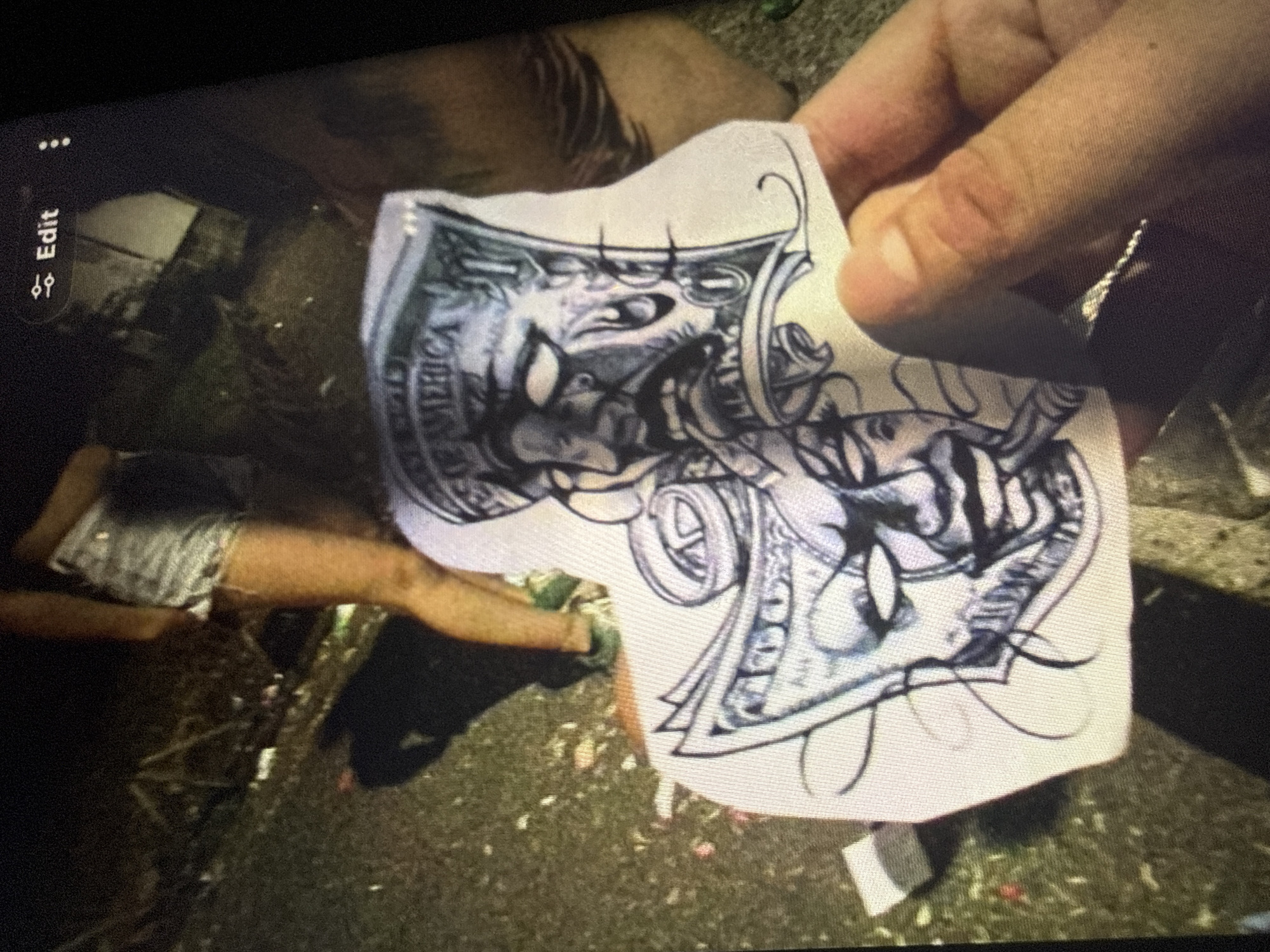 The image shows a close-up of a person's hand holding a paper stencil, likely for a tattoo. The stencil depicts two masked faces, one with a cigar in its mouth and the other with tears. Both faces are designed to resemble dollar bills, with "OF AMERICA" visible on one. In the background, out of focus, is a person wearing denim shorts and standing on a grassy, uneven surface littered with debris. The lighting suggests it is nighttime. There is no visible indication of the city of Sofia, Bulgaria.