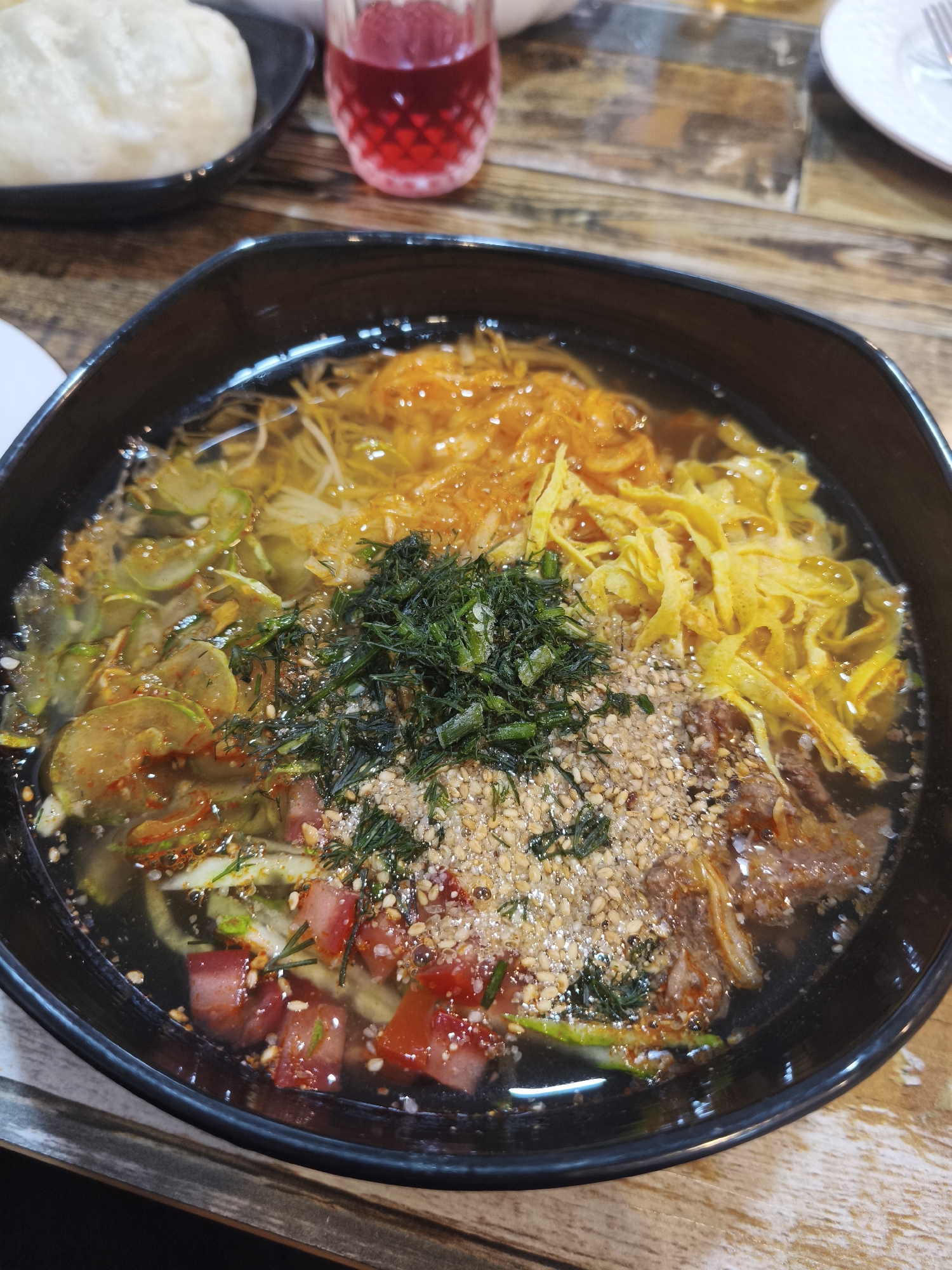 A close-up, overhead shot shows a black bowl of noodle soup on a wooden table. The soup contains a variety of ingredients, including shredded egg, thinly sliced green vegetables, chopped tomatoes, meat, and sesame seeds. A small dish with a white bun and a glass of red liquid are visible in the background. The scene is likely in Asan-si, South Korea.