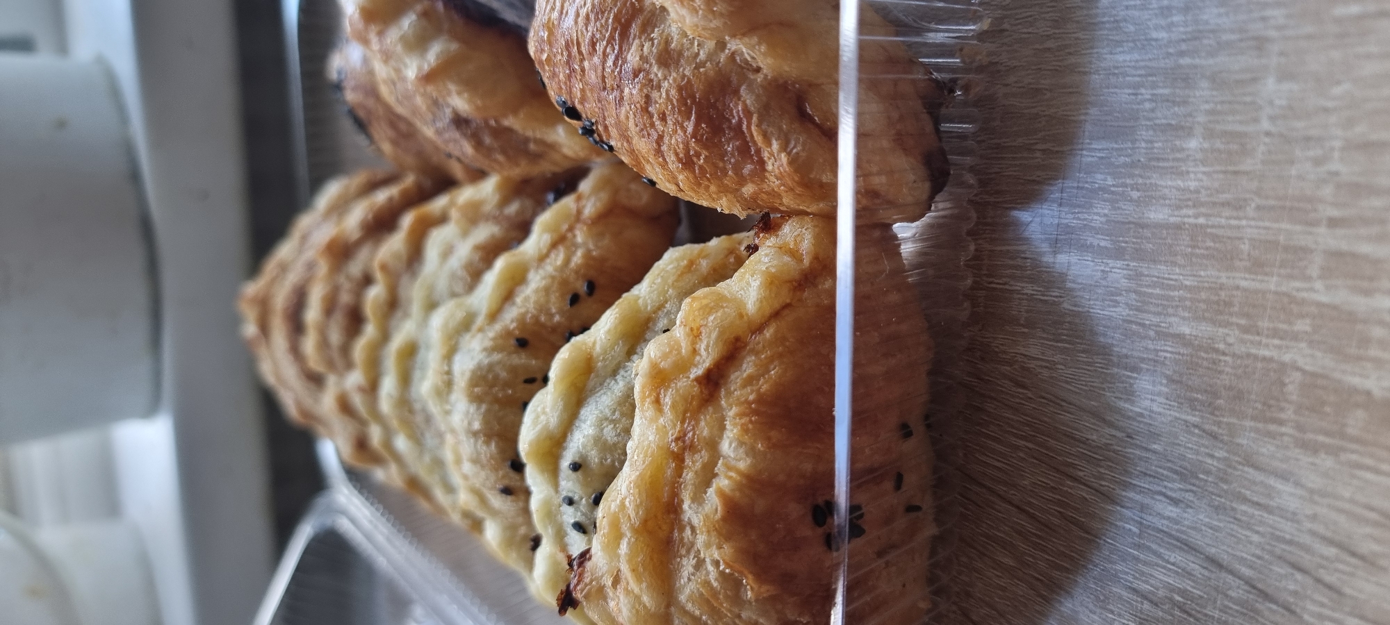The image shows a close-up of several baked goods, likely pastries or empanadas, arranged in a clear plastic container. The pastries are golden brown with visible flaky layers and are topped with black sesame seeds. They are positioned on a surface that appears to be a wooden table or countertop. The background is out of focus, showing a white surface, possibly a windowsill or shelf. The perspective is from above and slightly to the side, focusing on the texture and details of the baked goods. There is no information in the image to suggest a specific location in Aktobe, Kazakhstan.