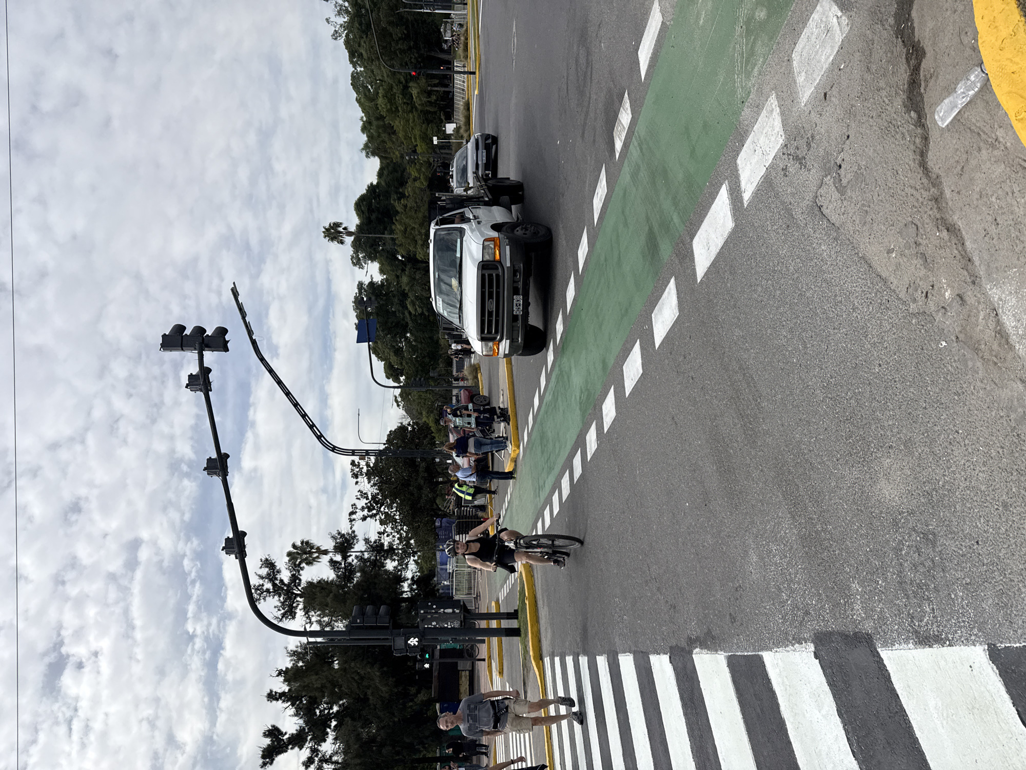 The image shows a street scene in Buenos Aires, Argentina, featuring a marked bicycle lane. A cyclist rides across a crosswalk adjacent to the bike lane. A white pickup truck and a dark sedan are stopped on the street. Pedestrians are on the sidewalks. Traffic lights are visible. The sky is overcast.
