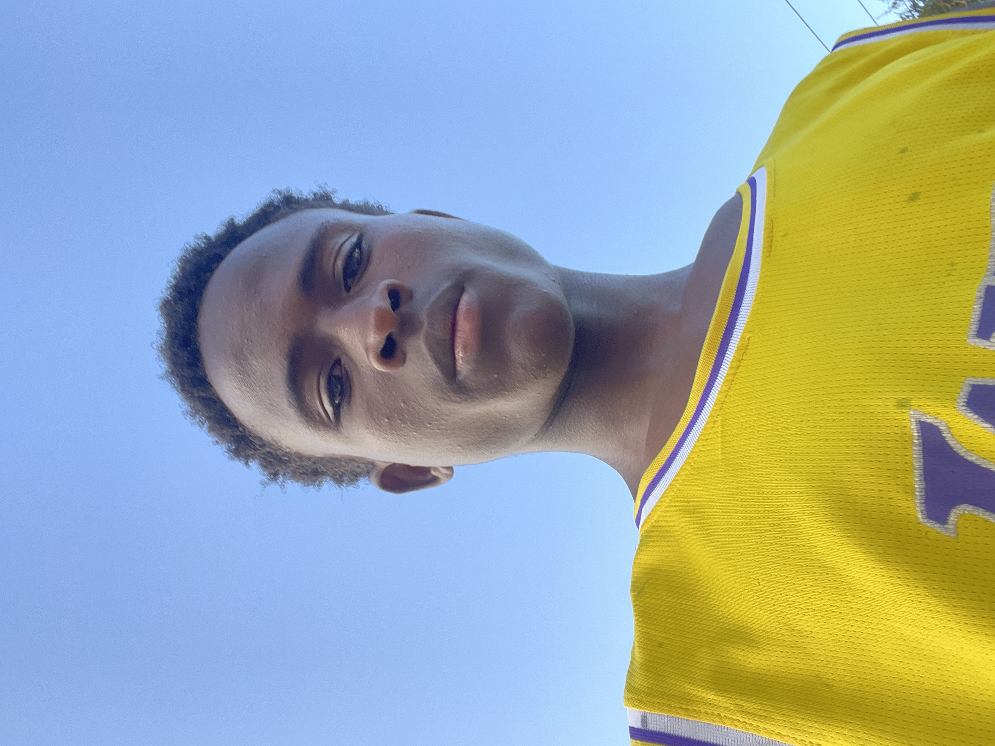 A low-angle, close-up, waist-up outdoor shot features a young Black male against a clear, bright blue sky. The subject is positioned centrally, looking directly at the camera with a neutral expression. He has dark, curly hair and his skin is a deep brown tone, with visible pores and some minor blemishes on his face. He is wearing a yellow basketball jersey with purple and white trim around the neckline and armholes. The jersey has a large purple "L.A." logo visible on the lower left side of the frame. The lighting suggests it is daytime, possibly late afternoon or early morning due to the soft quality of the light. The background is entirely composed of the sky, with no discernible features to indicate the specific location within Jos, Nigeria, beyond it being an outdoor urban or suburban setting. There are no other people or objects prominently featured in the frame.